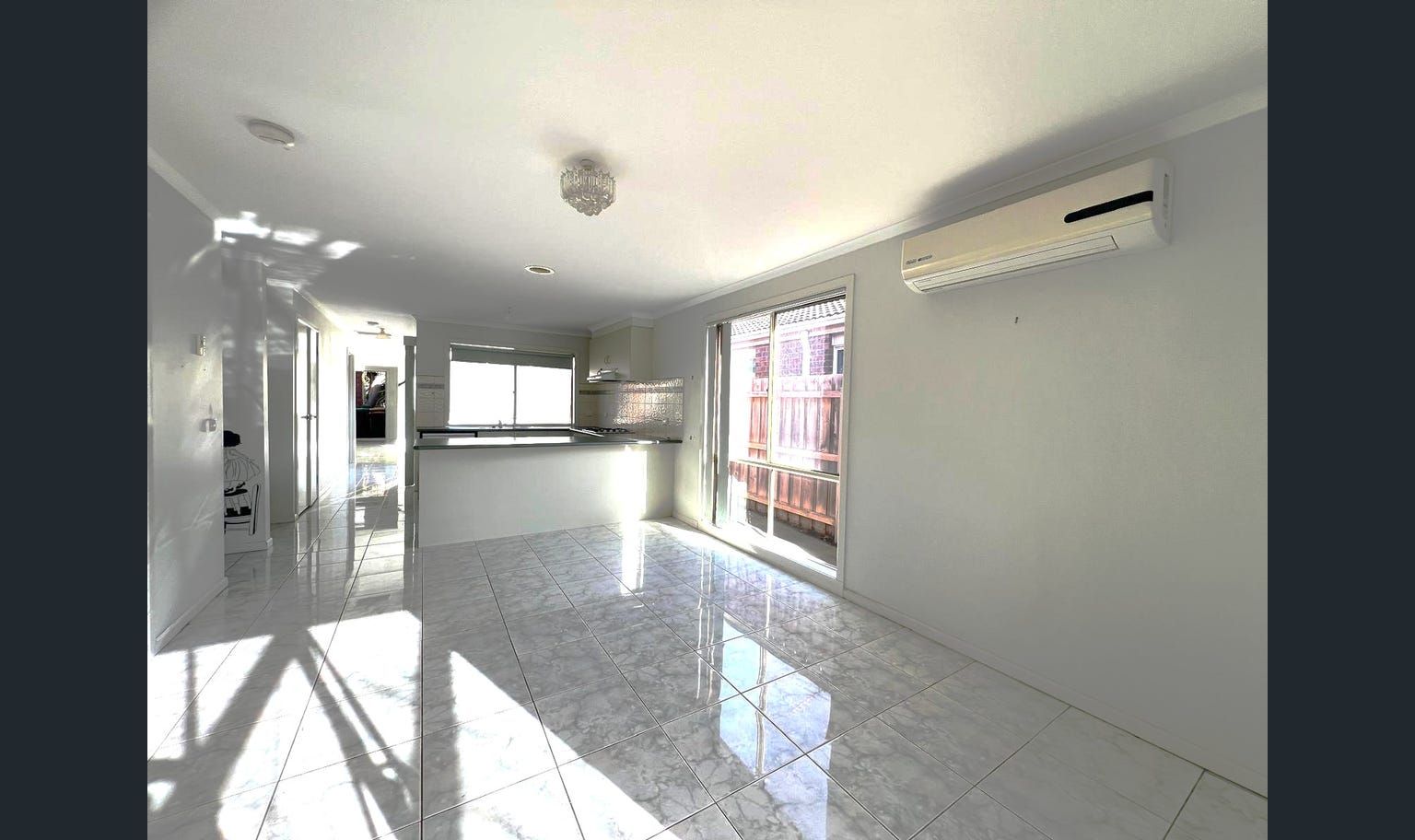 Bright, empty living space with white walls and tiled floor. Includes a kitchen, sliding door, and air conditioning unit.