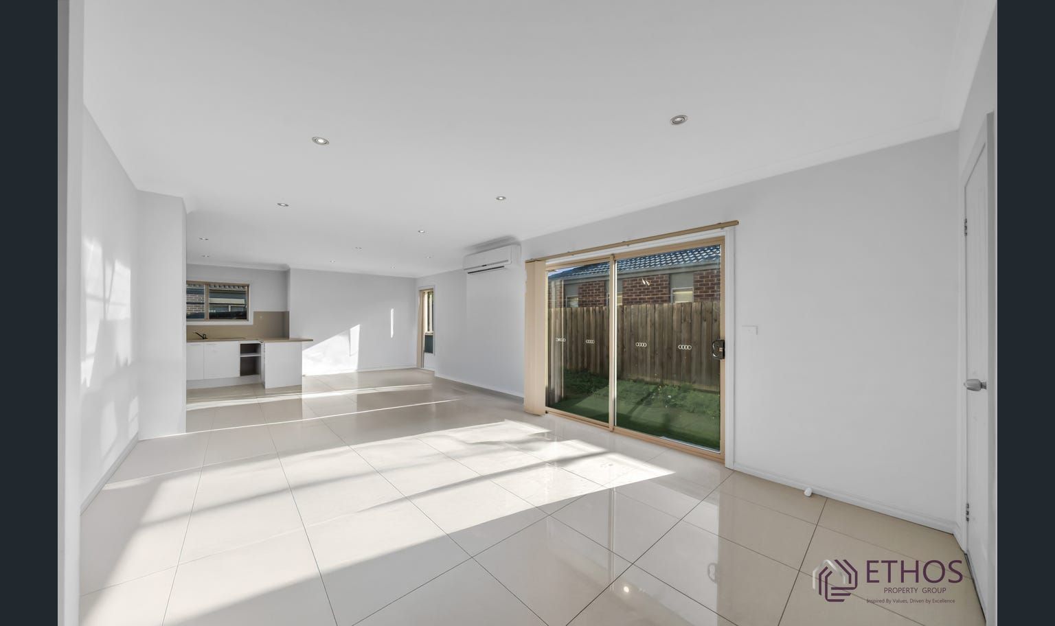 Empty, bright, modern living room with large sliding glass door. Kitchen with white cabinets visible in background.