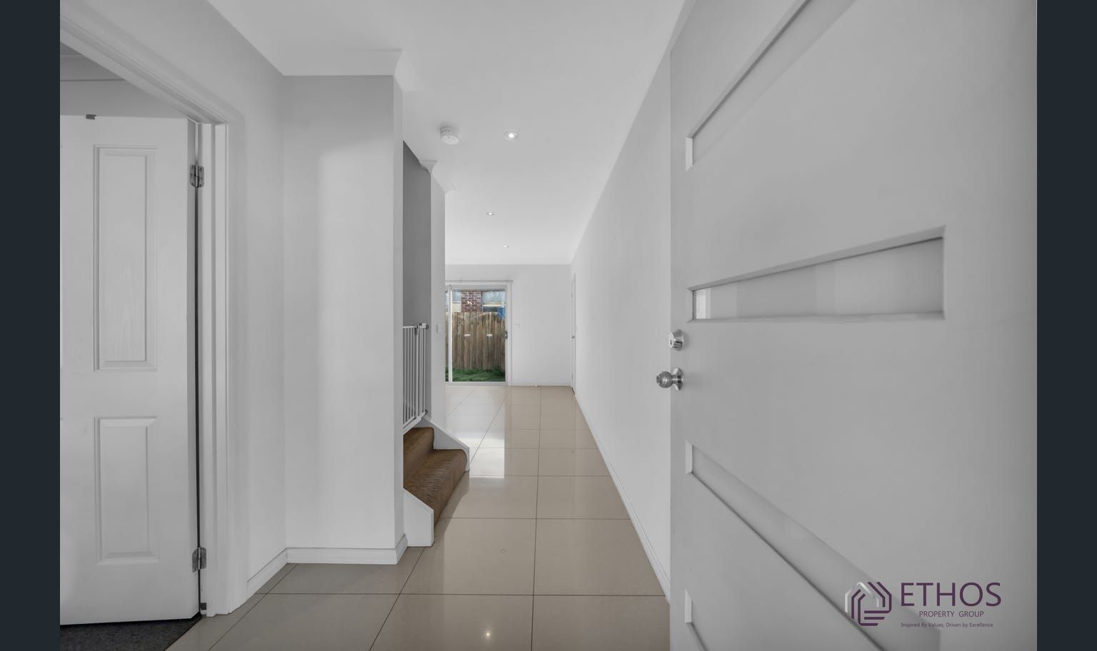 Hallway with tiled floor, white walls and doors, leading to a backyard view.