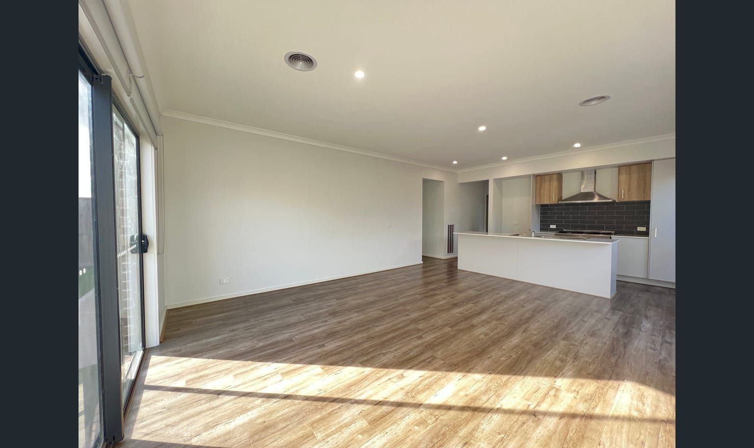 Empty, modern living space with wood flooring, open to a kitchen with light cabinets.