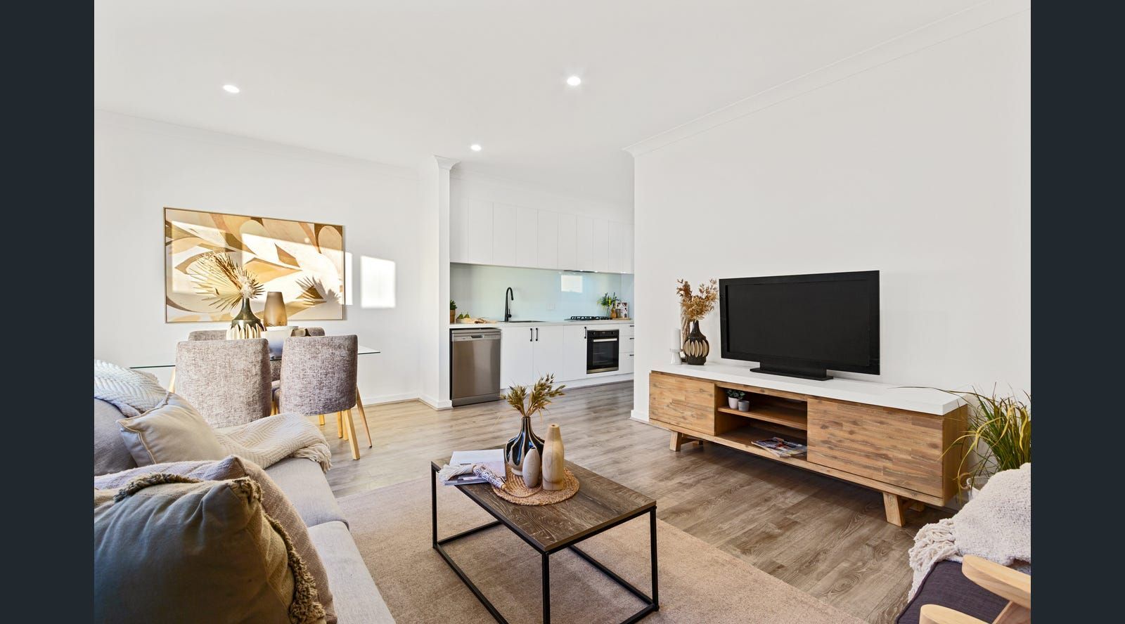 Bright living room with white walls, hardwood floors, and a kitchen in the background.