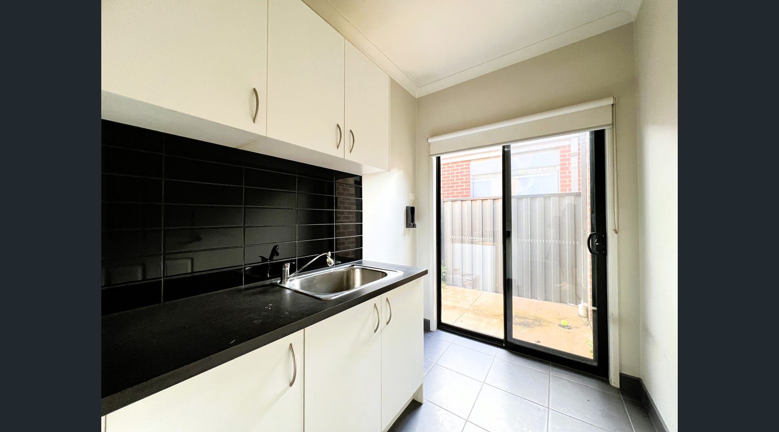 Laundry room with white cabinets, black countertop, stainless steel sink, black tiled backsplash, and sliding glass door.