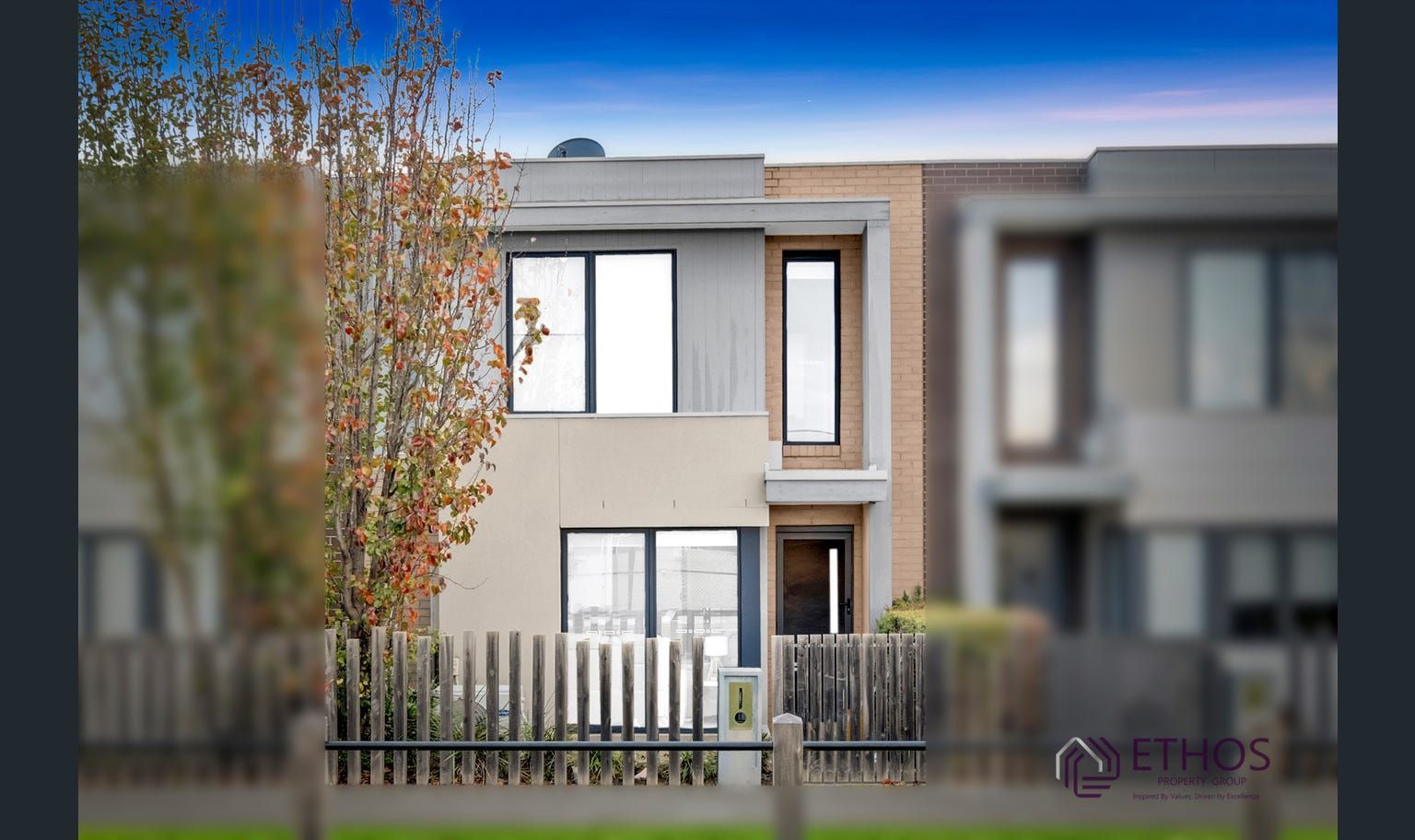 Modern two-story townhouse with neutral exterior, light-colored fence, and small tree in front.