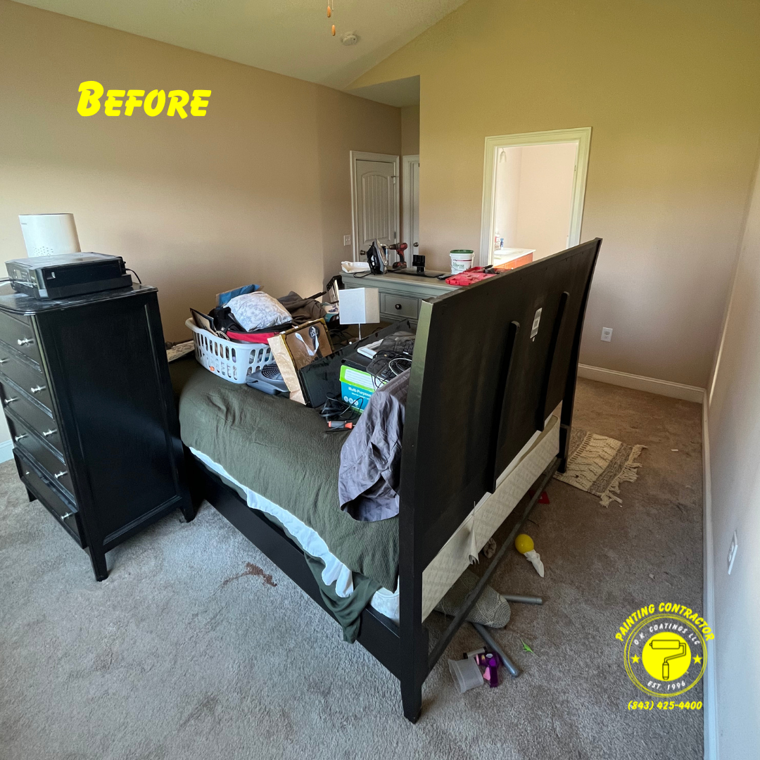 A beige bedroom before renovations featuring a black dresser, a bed with a dark frame, and clutter on the bed and floor.