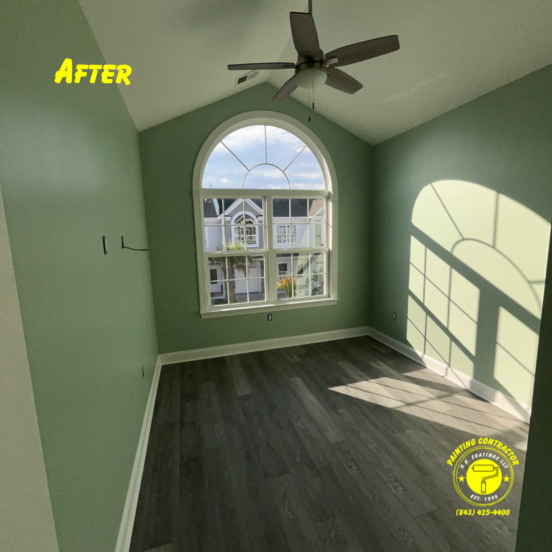 A room with sage green walls, dark wood floors, a vaulted ceiling with a fan, and a large arched window.