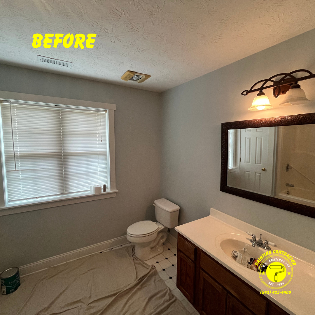 A bathroom with light grey walls, a white vanity, a toilet, and a window covered by blinds before a renovation.