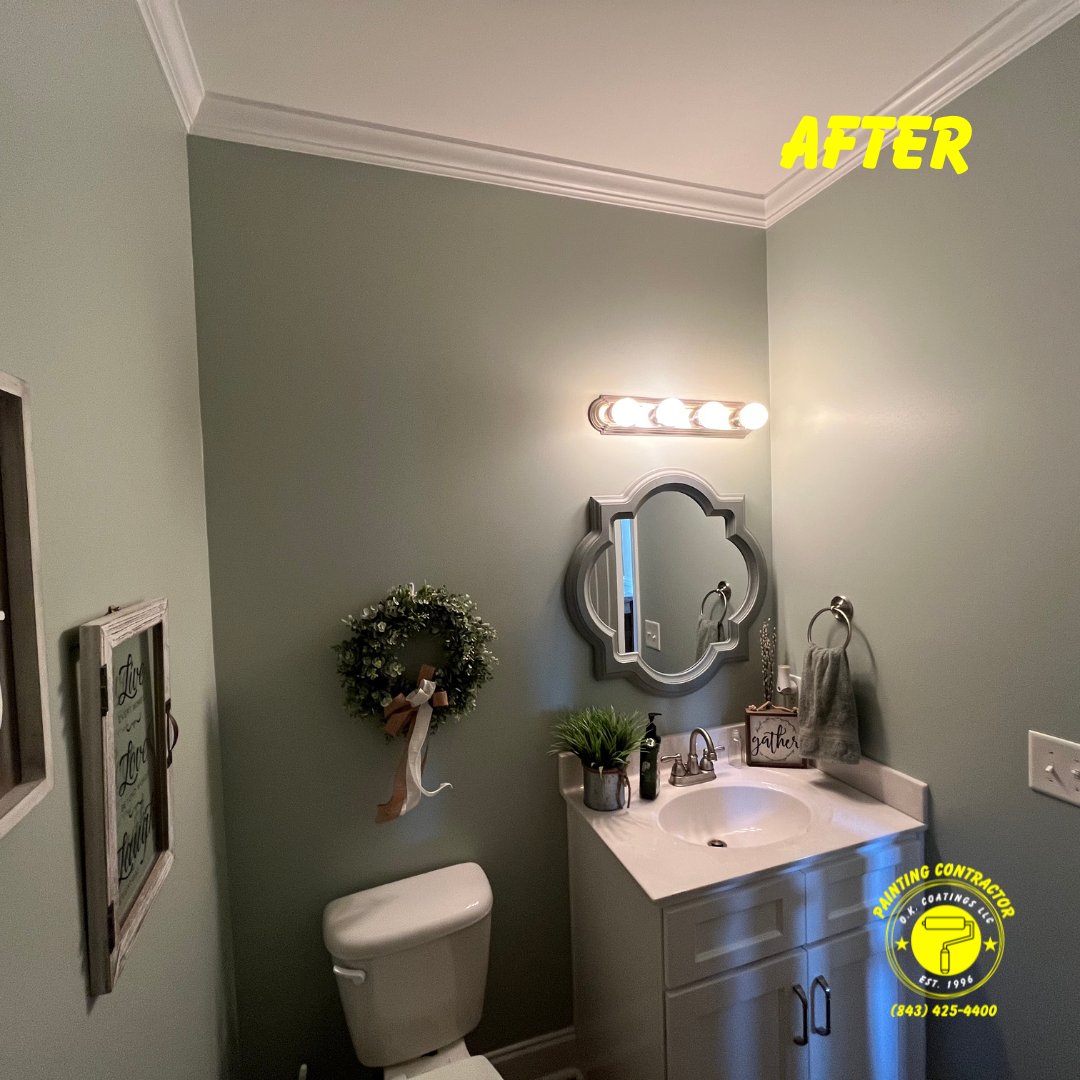 A bathroom painted in soft sage green with a white vanity, a decorative mirror, and a floral wreath hanging on the wall.