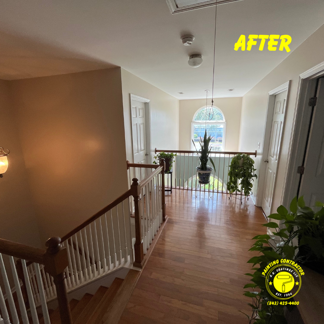 An interior hallway with hardwood floors, a staircase with white railings, and potted plants near a windowed landing.