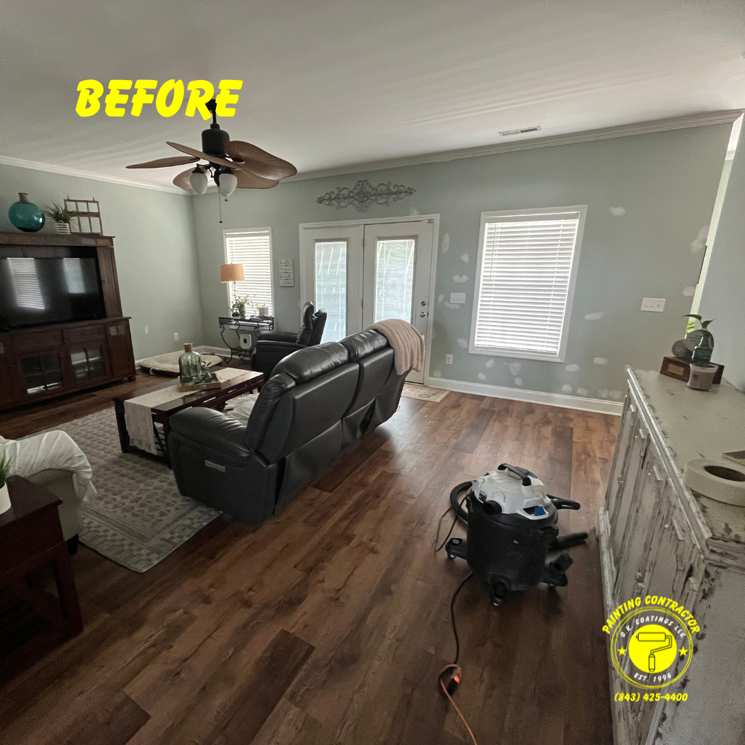 A living room before painting, featuring grey walls with patch marks, a dark leather sofa, wood floors, and a shop vacuum.