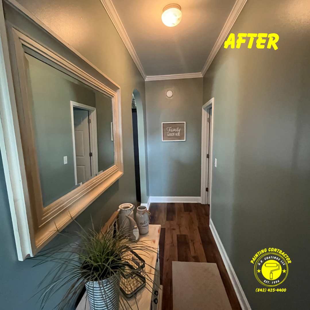 A hallway with sage green walls, wood flooring, a large mirror, and a small table with a potted plant under a ceiling light.