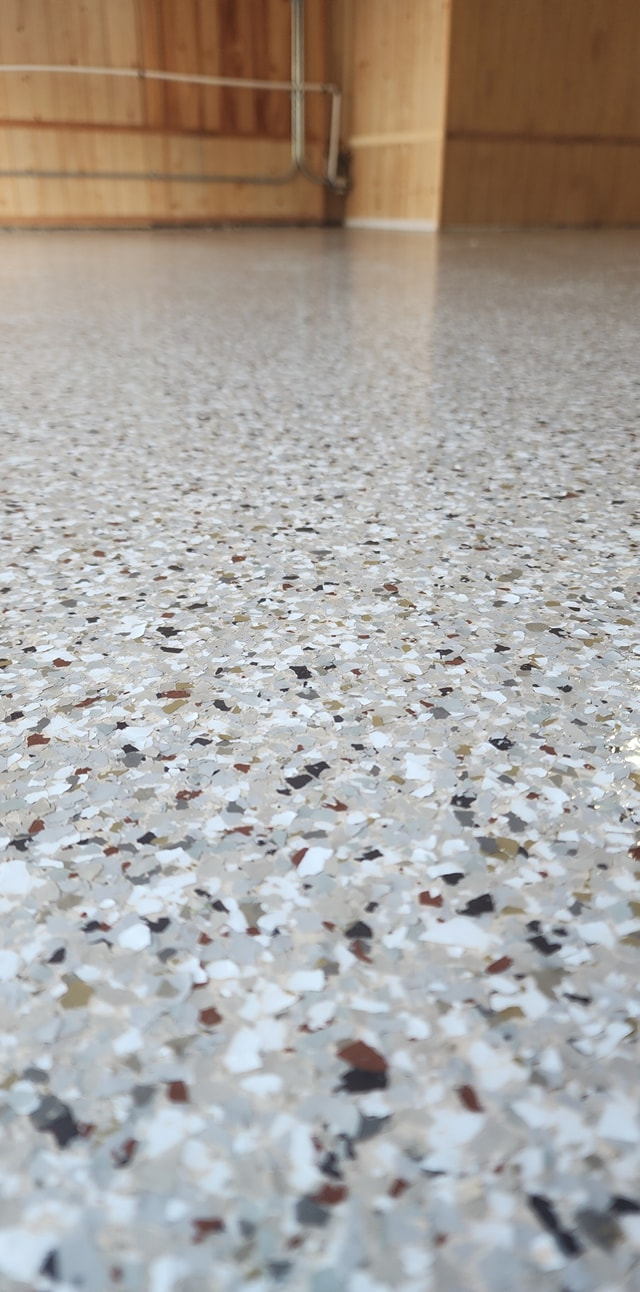 Close-up of a speckled floor with light gray, white, and dark brown flecks, with wood paneling and a pipe in the background.