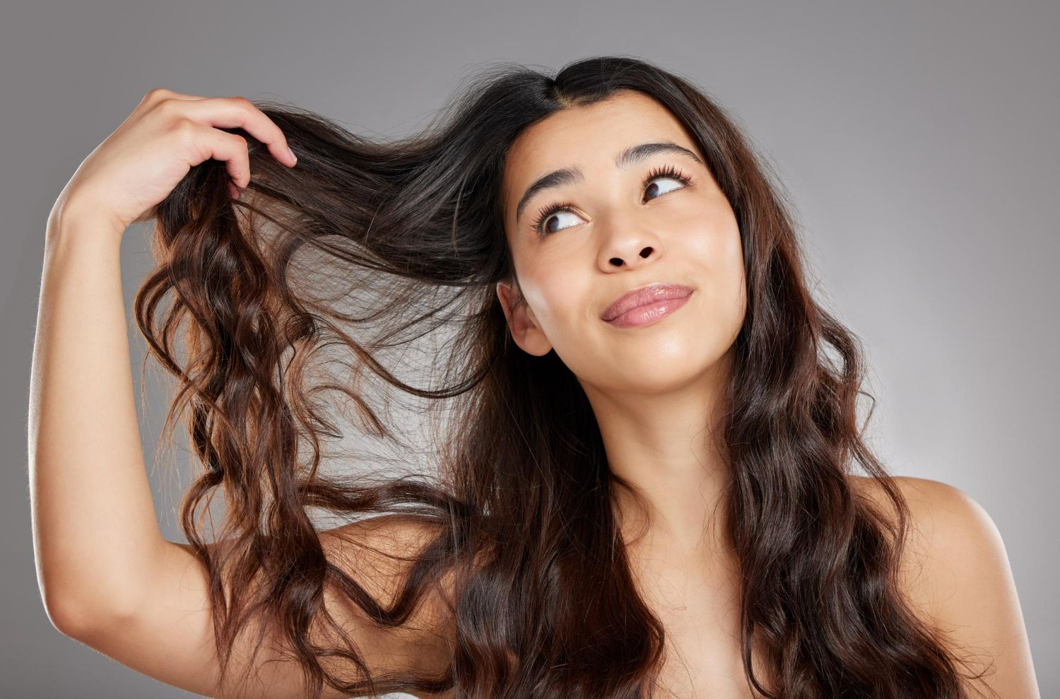 A person with long, wavy dark hair looks upward while holding a section of their hair to examine the texture.