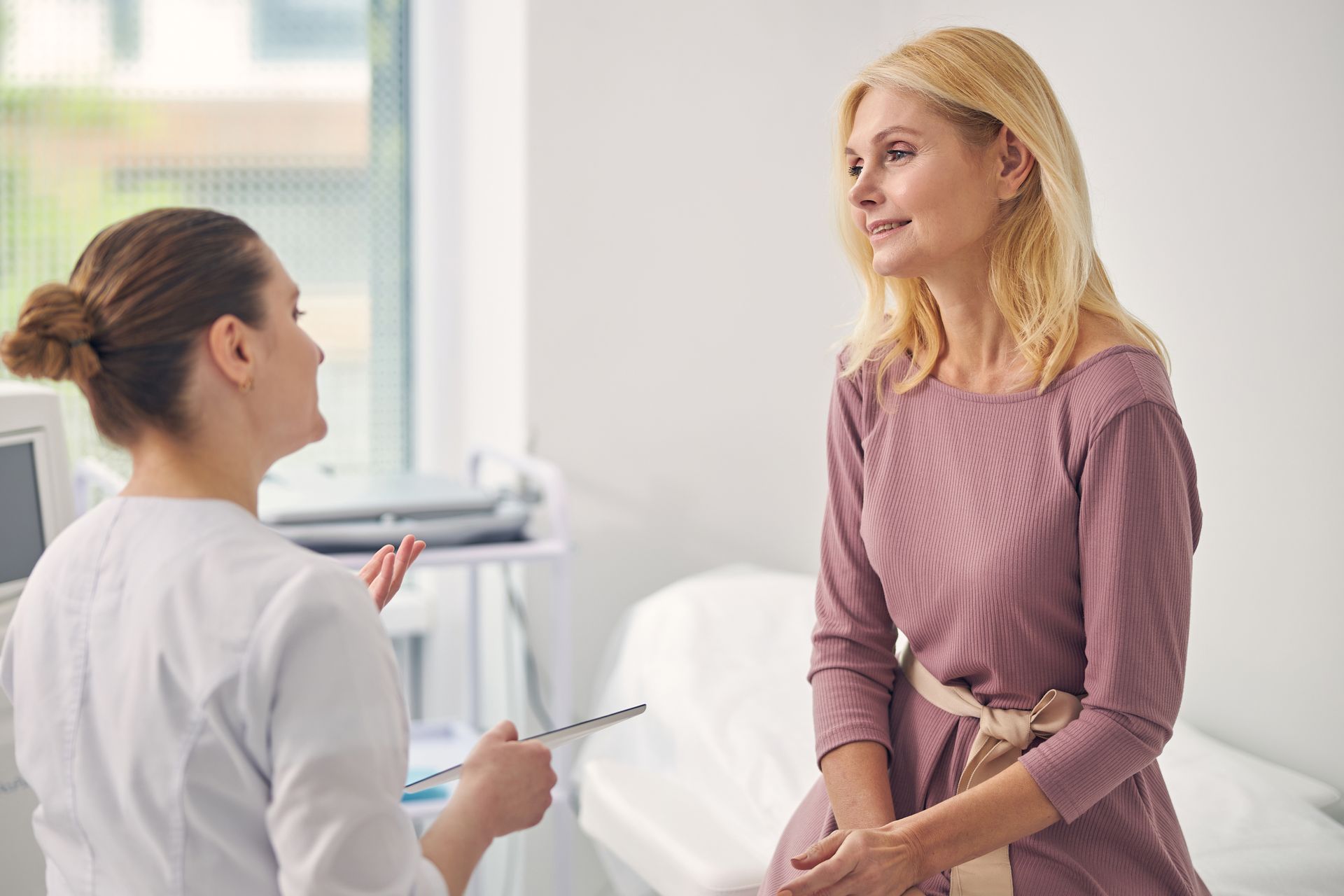 A healthcare provider in a white coat talks to a patient in a pink dress in a medical examination room.
