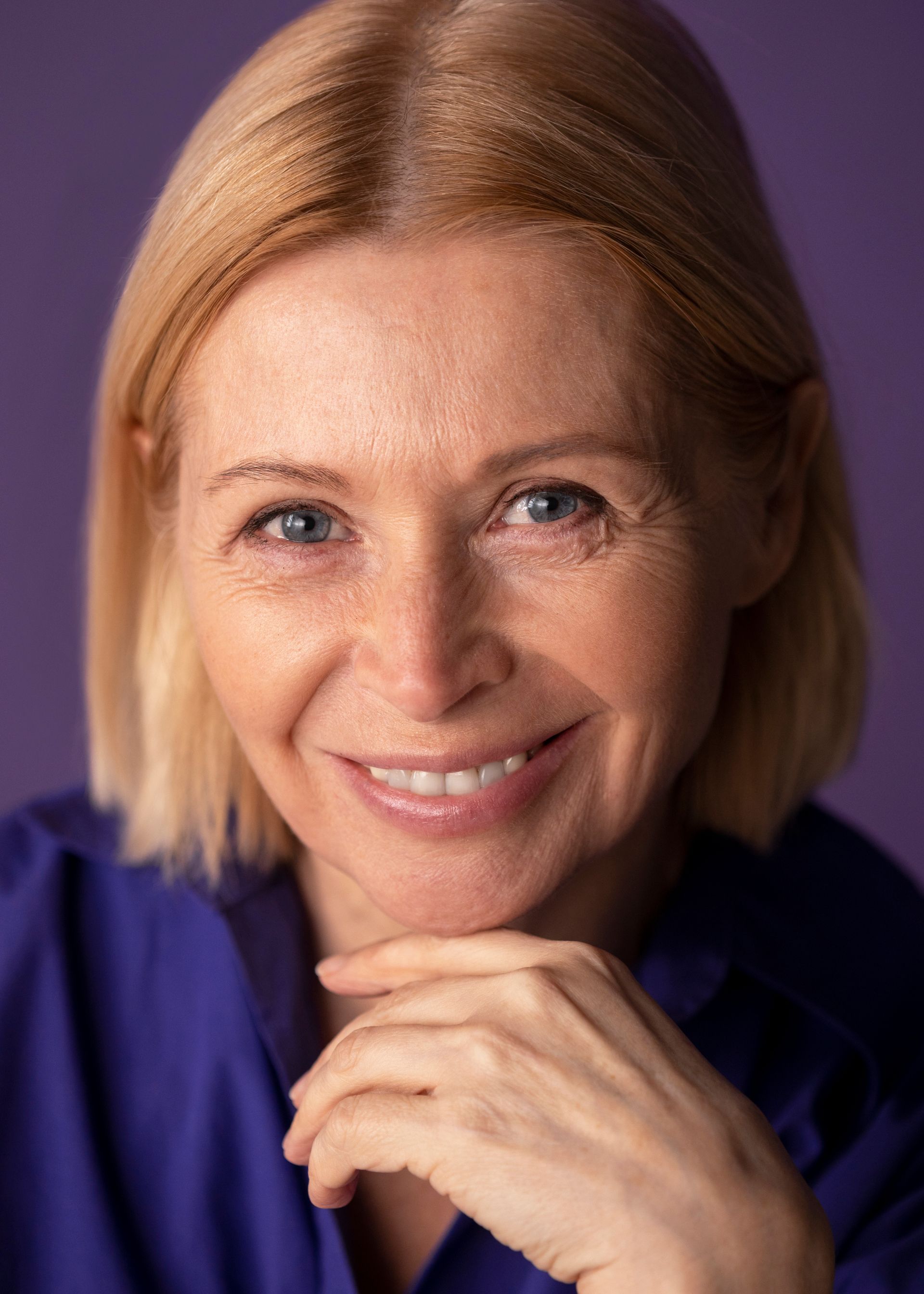 A woman in a purple shirt is smiling with her hand on her chin.