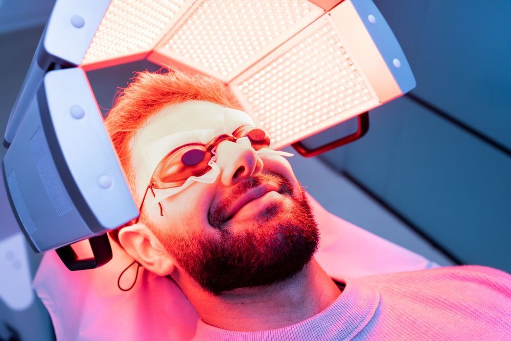 A person lies back with protective goggles while receiving red light therapy from an overhead LED panel.