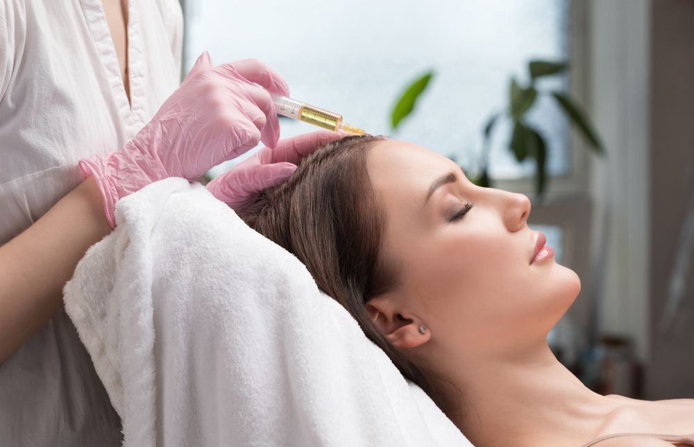 A professional in pink medical gloves performs a scalp injection treatment on a client lying on a clinic bed.