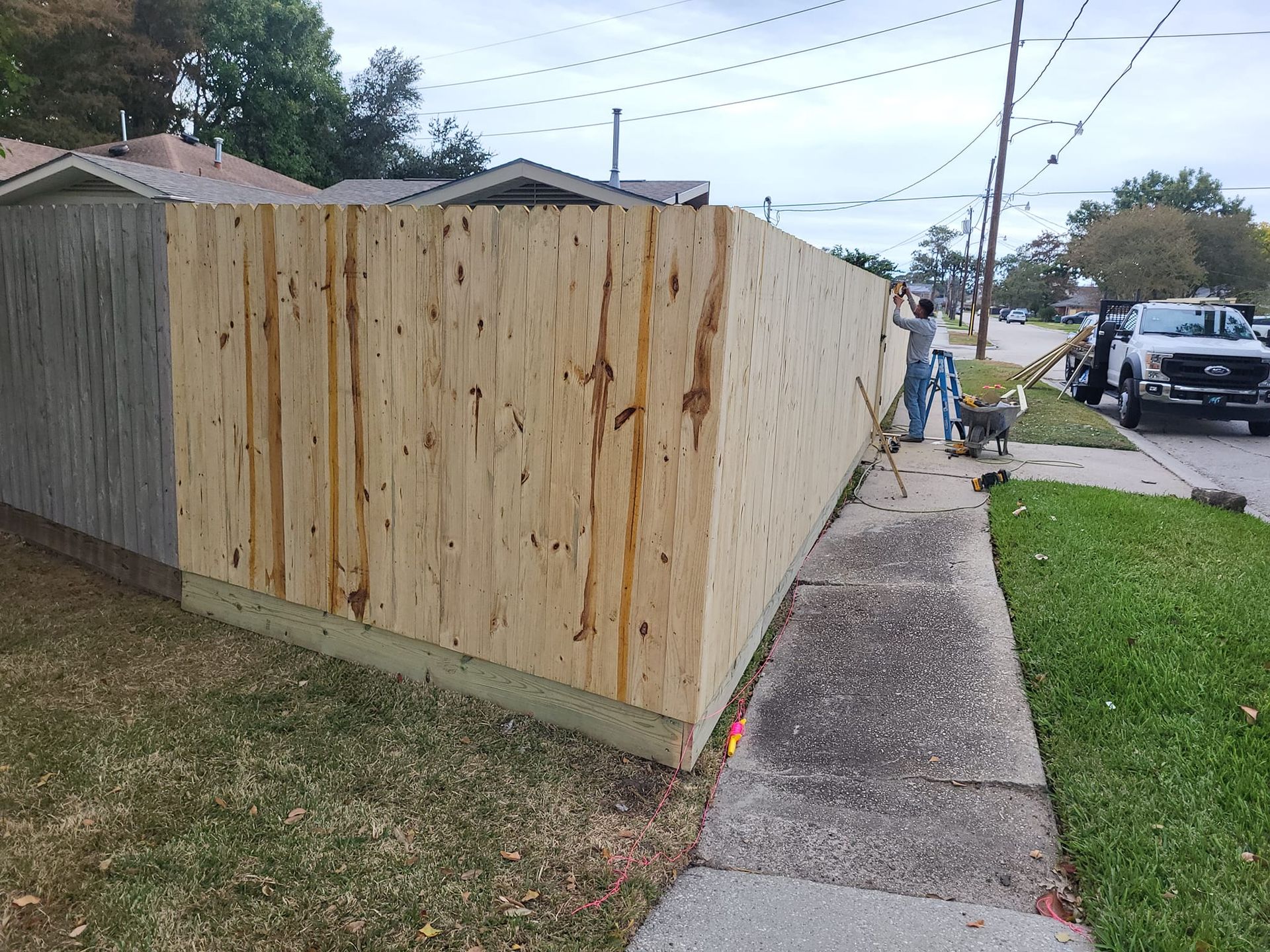 A man is standing next to a wooden fence on the sidewalk.
