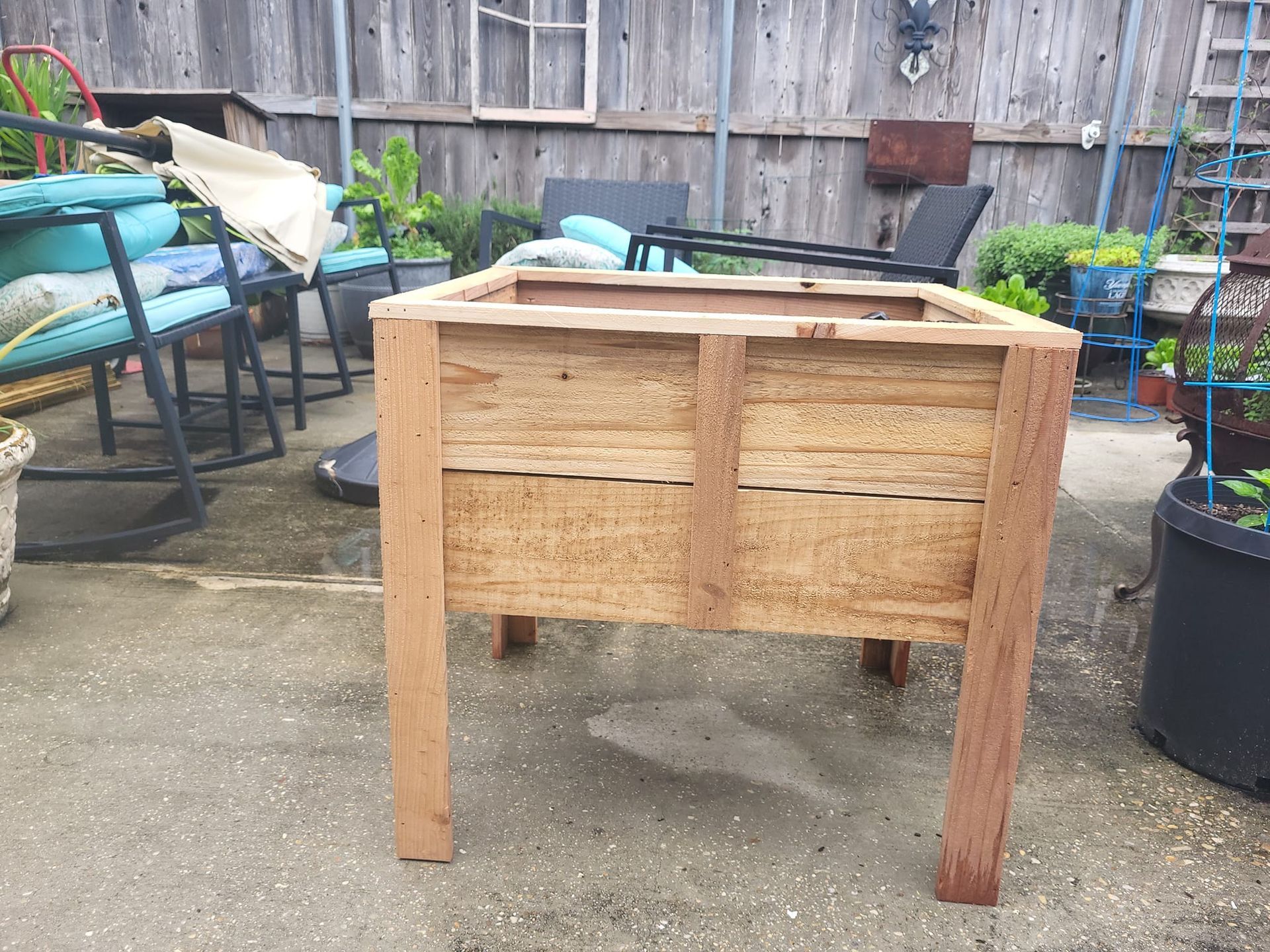 A wooden planter is sitting on a patio next to rocking chairs and potted plants.