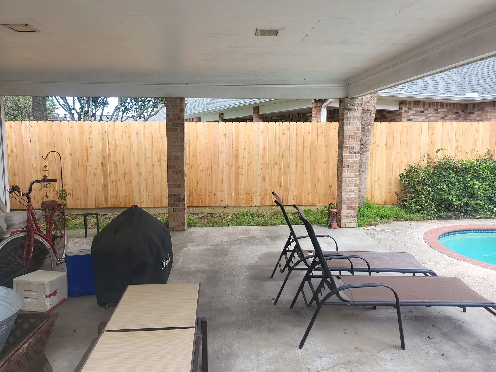 A patio with a wooden fence , chairs , tables and a pool.