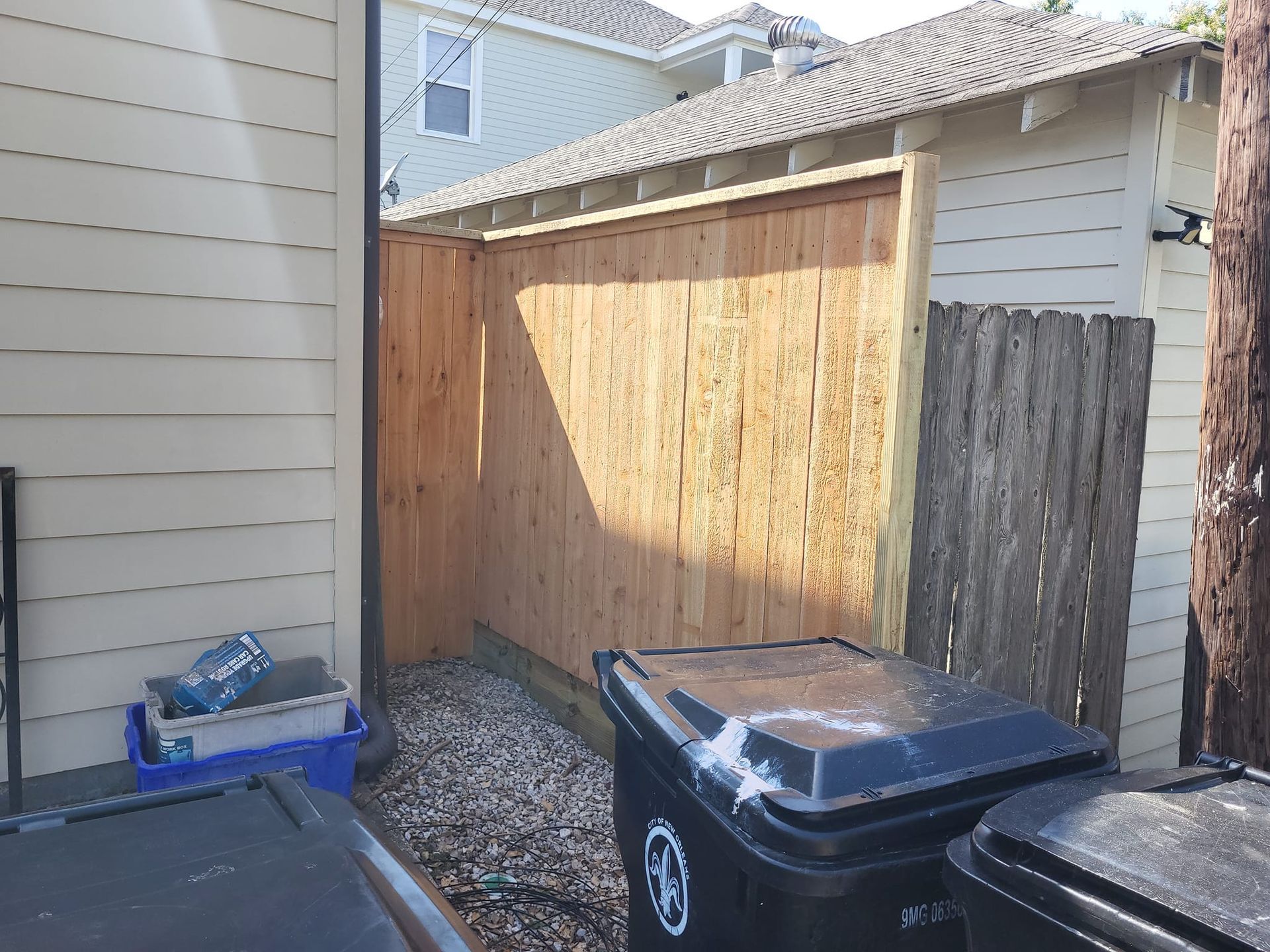 A wooden fence is surrounded by trash cans in a backyard.