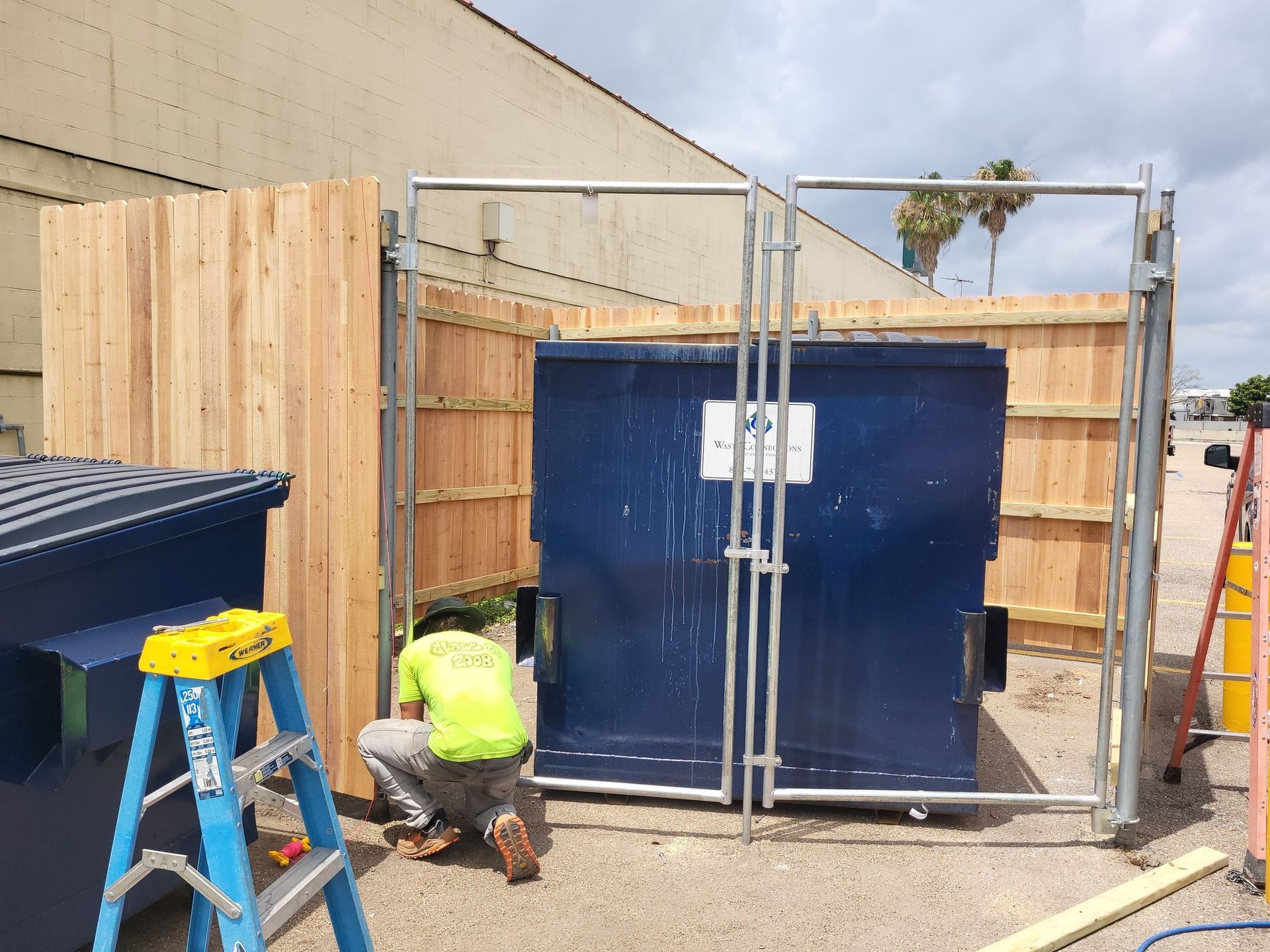 A man is working on a wooden fence around a dumpster.