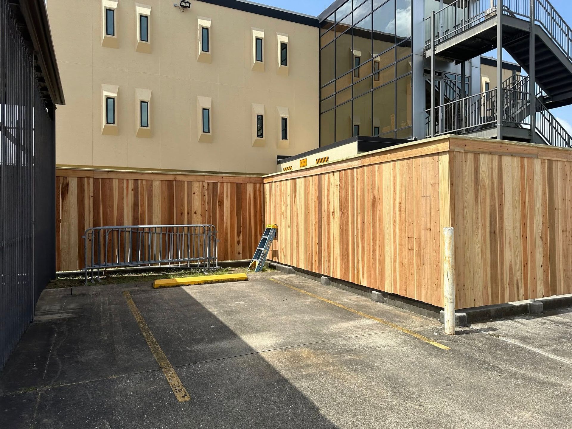 Wooden fence in a parking lot, with a building in the background. A ladder stands near the fence.
