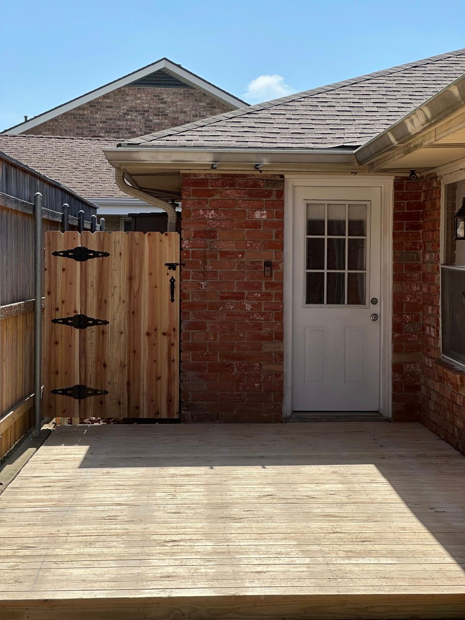 Wooden deck with gate, brick wall, and white door, under a rooftop on a sunny day.
