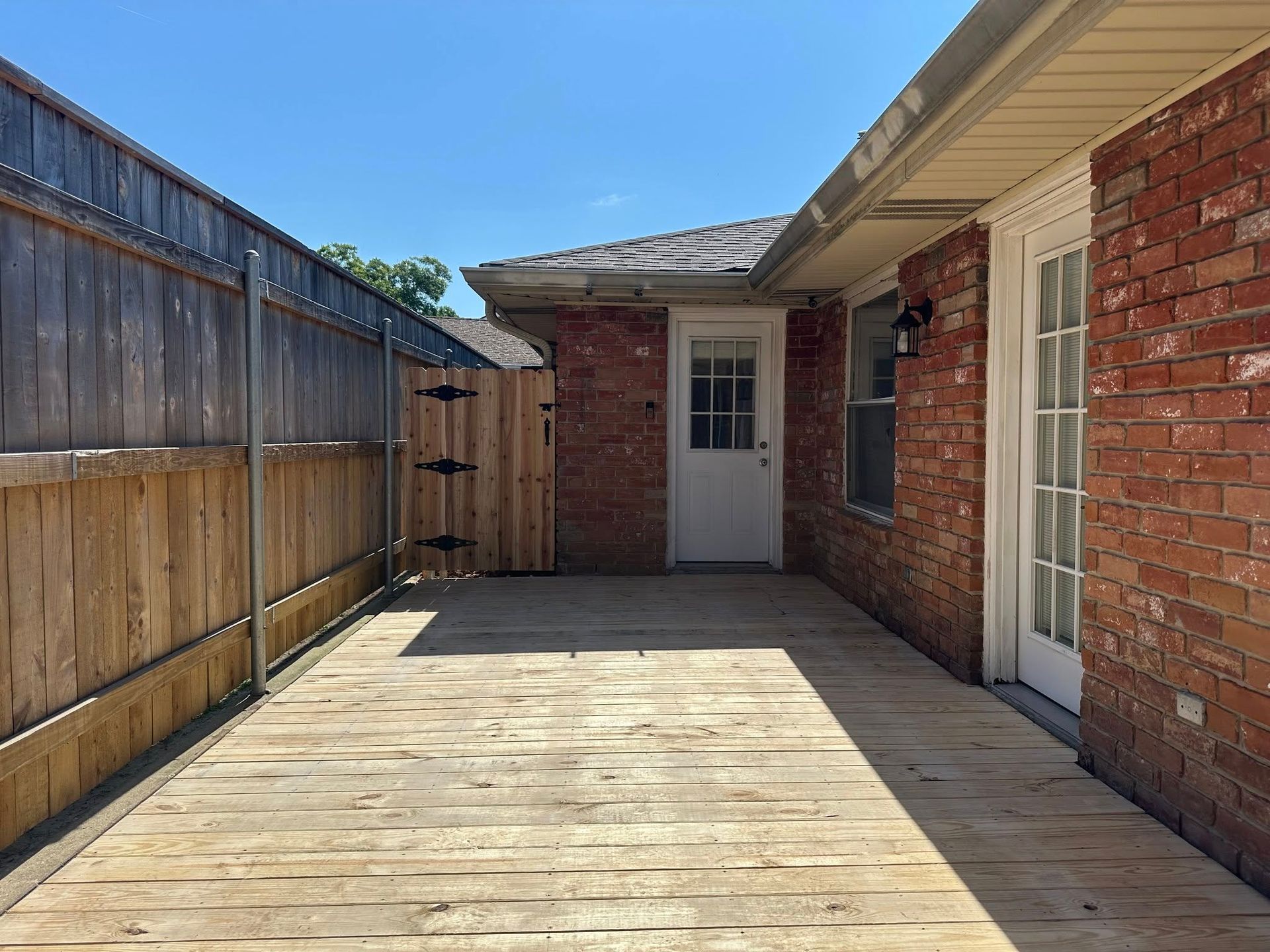 Wooden deck in a backyard. Brick wall, wooden fence, and white door. Bright, sunny day.