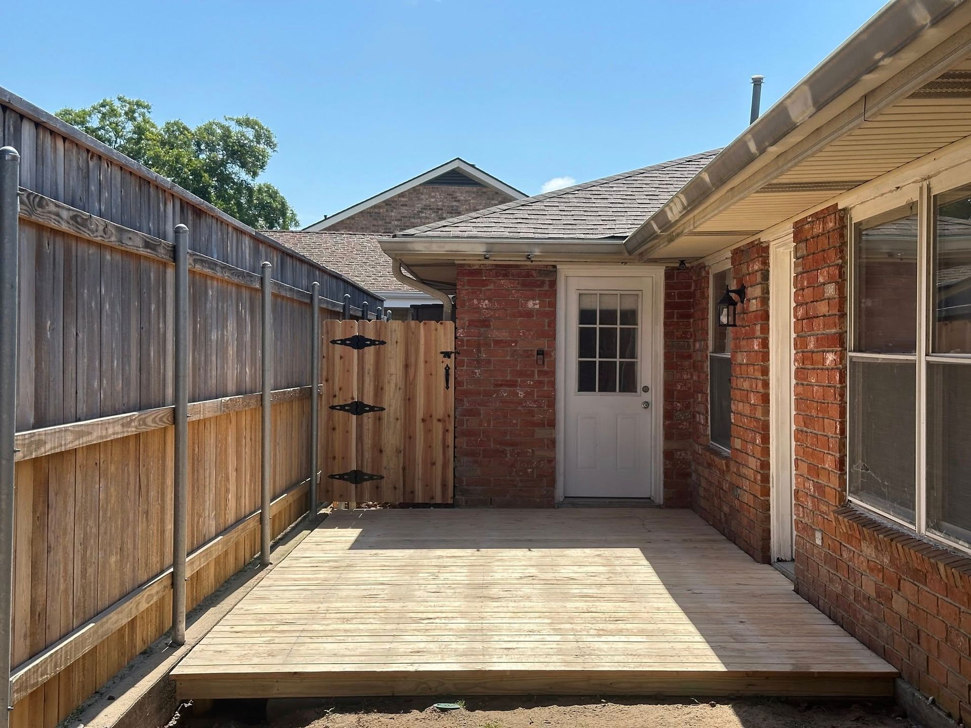 Wooden deck in a backyard, enclosed by fences and a brick wall, with a door and blue sky.
