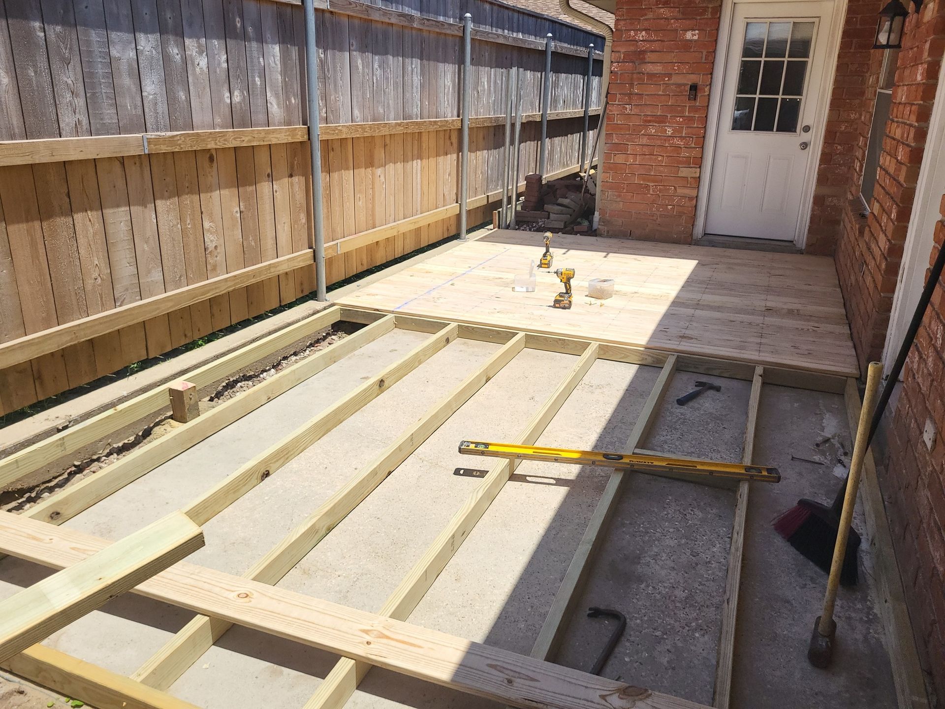 Deck frame construction in a backyard, with a partially built wooden structure against a fence and brick wall.