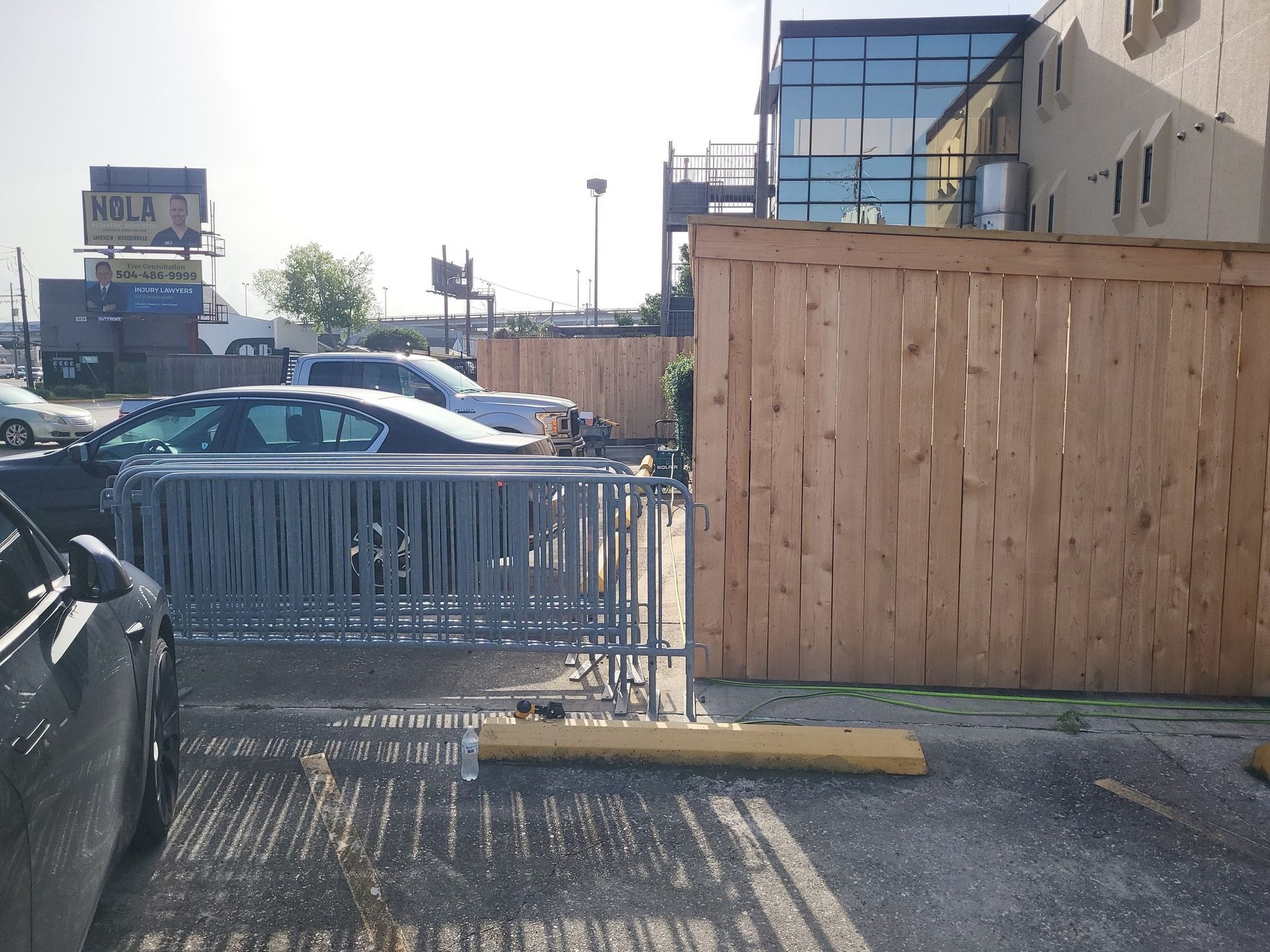 Cars parked in a parking lot next to a wooden fence and metal barricade. A building is in the background.