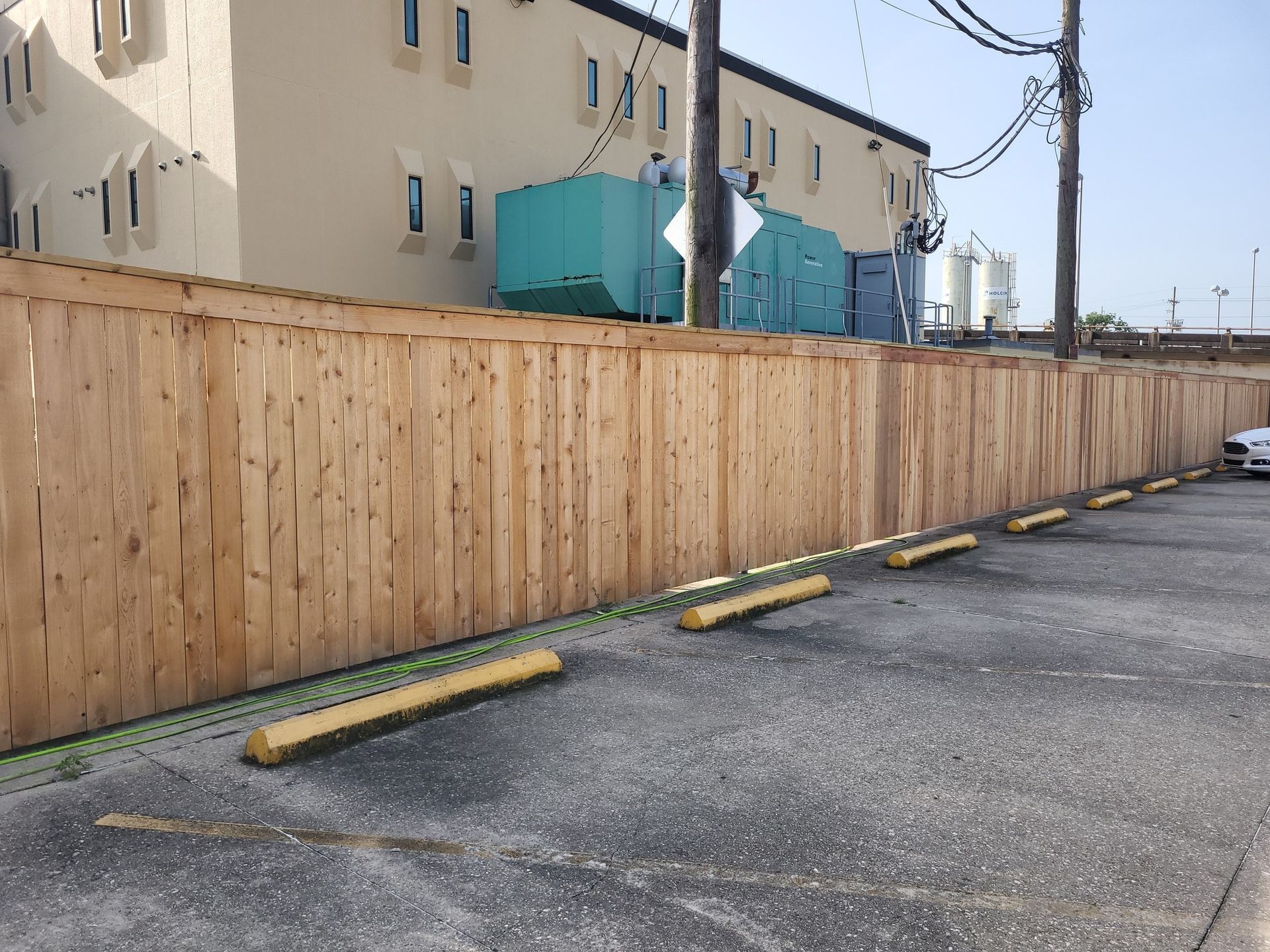 Wooden fence along parking lot, yellow parking bumpers, building in the background, sunny day.