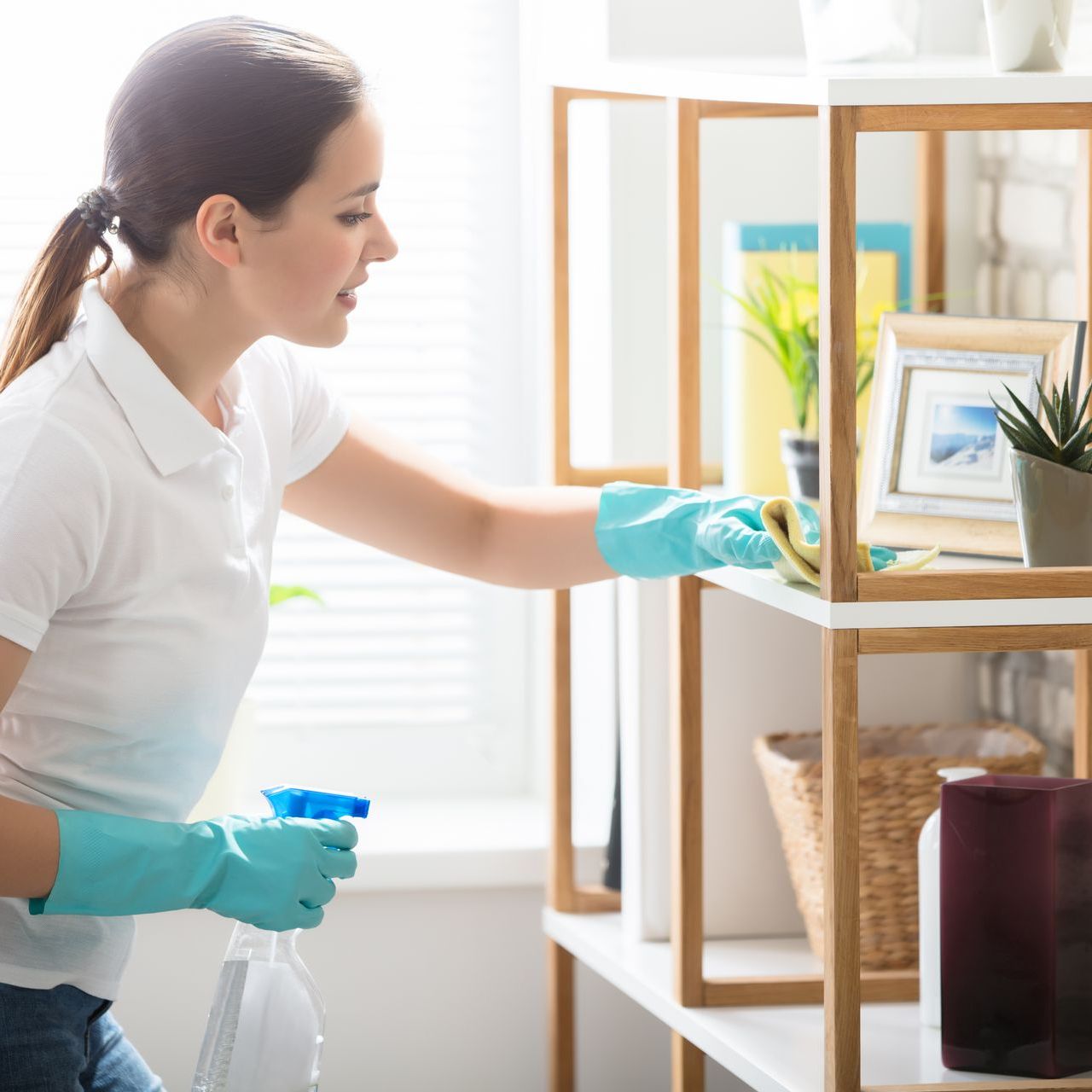 A woman is cleaning a shelf with a spray bottle and a cloth.