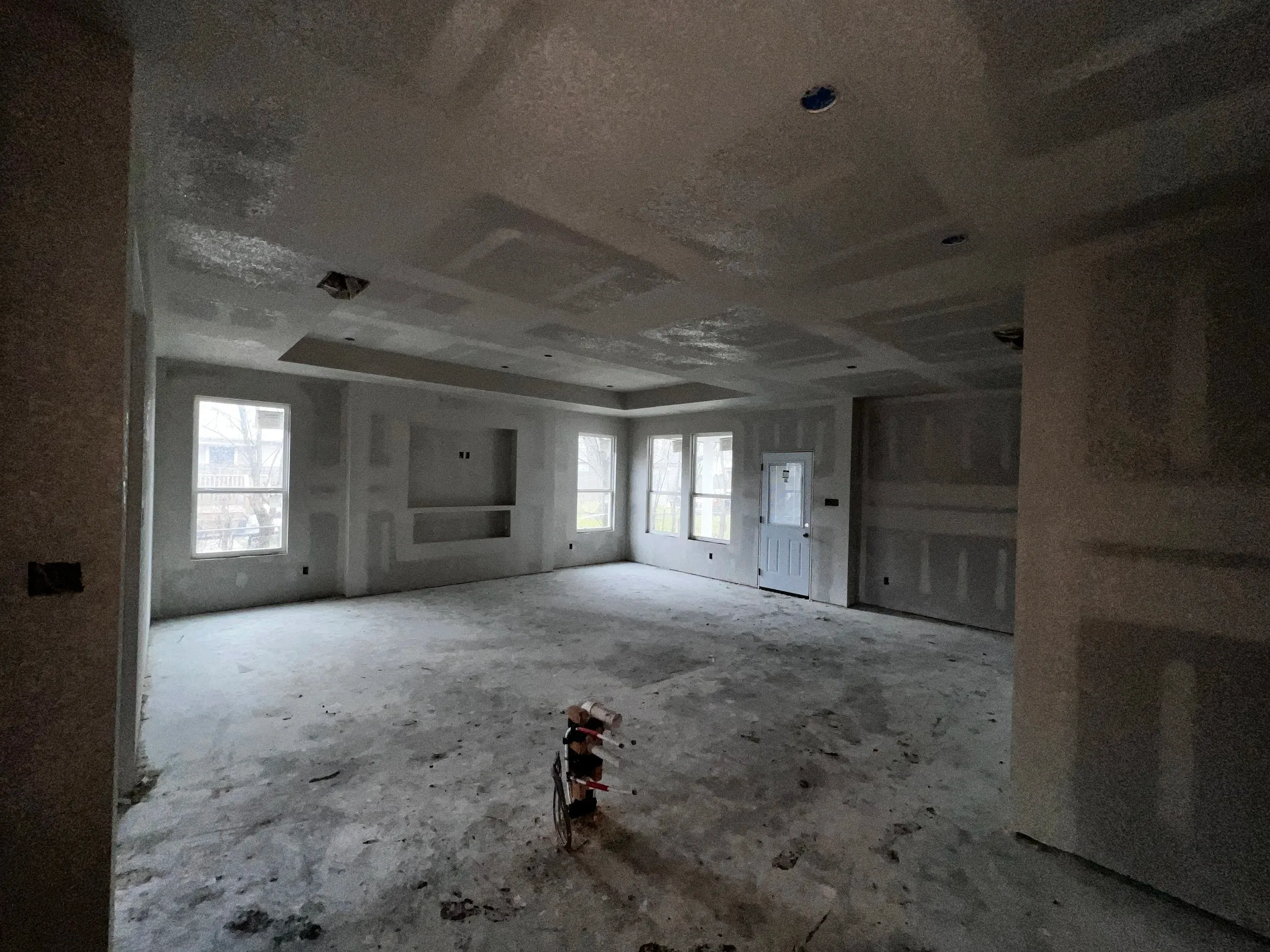 Empty unfinished room with exposed walls, windows, and dusty floor, lit by daylight.