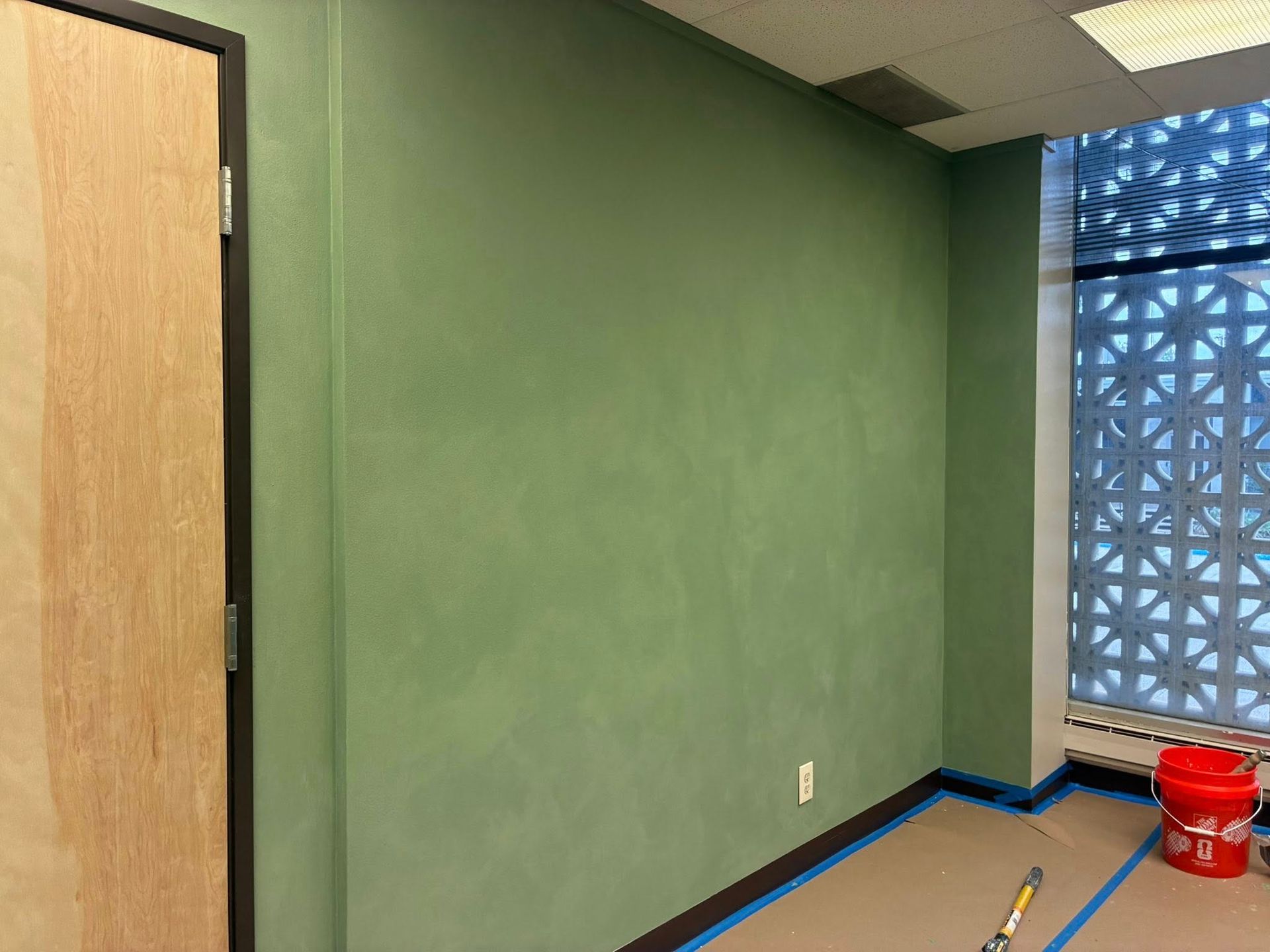 Empty room with green walls, wood door, blue-taped floor edges, and a red bucket by a window.