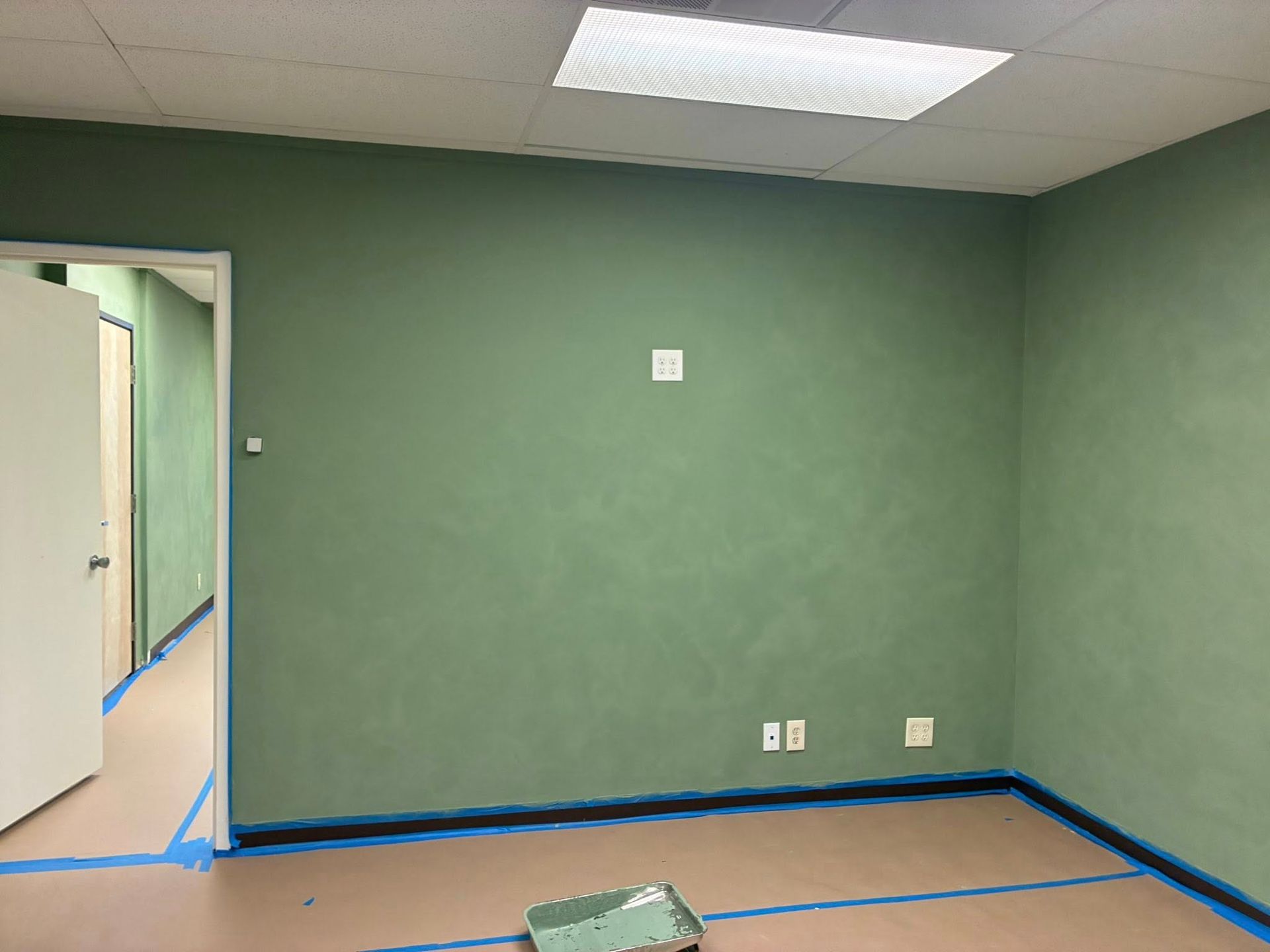 Empty green room with blue tape on the floor, open doorway, and ceiling lights.