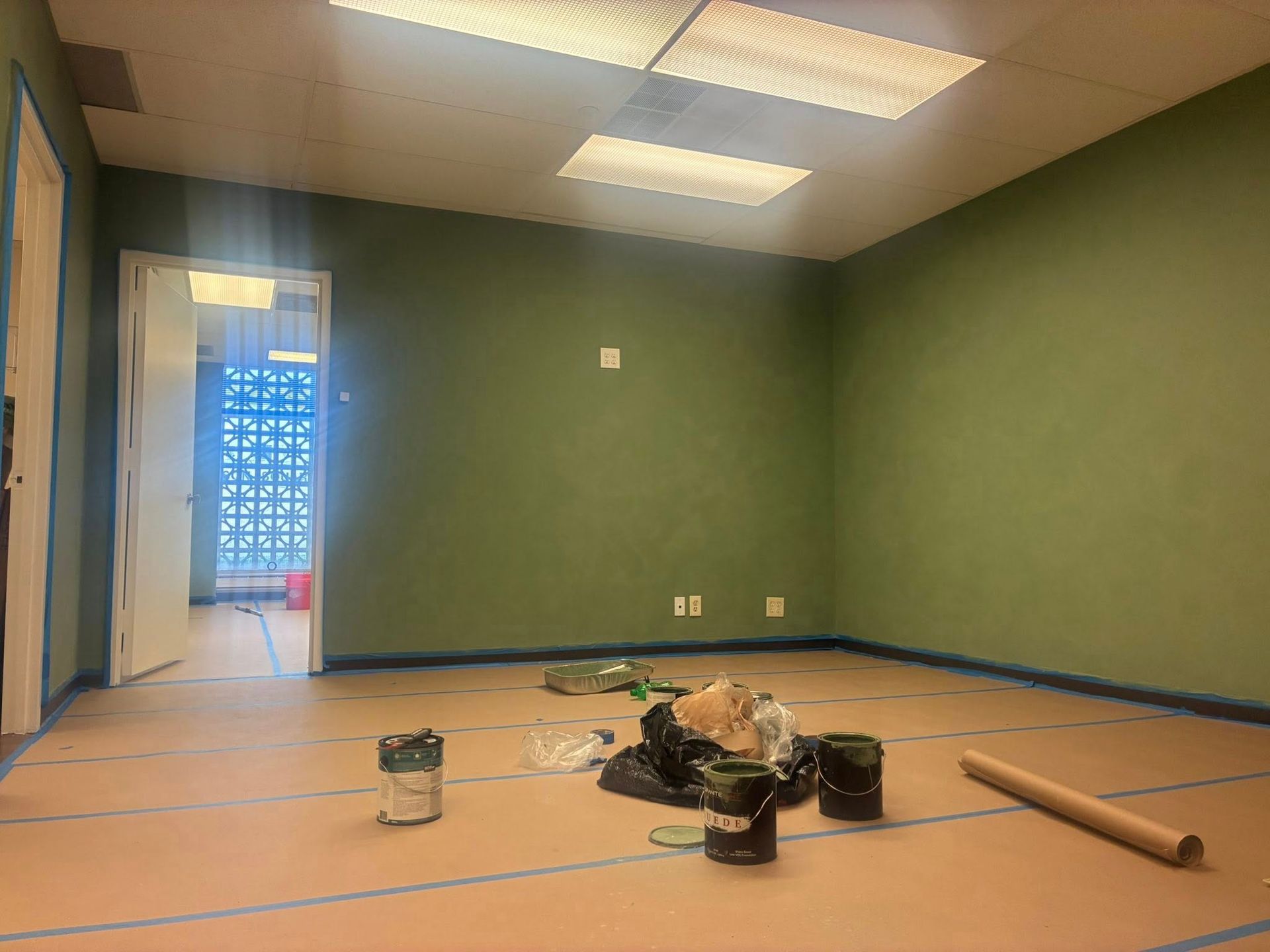 Empty green room with ceiling lights, paint supplies on the floor, and a doorway to a tiled hall