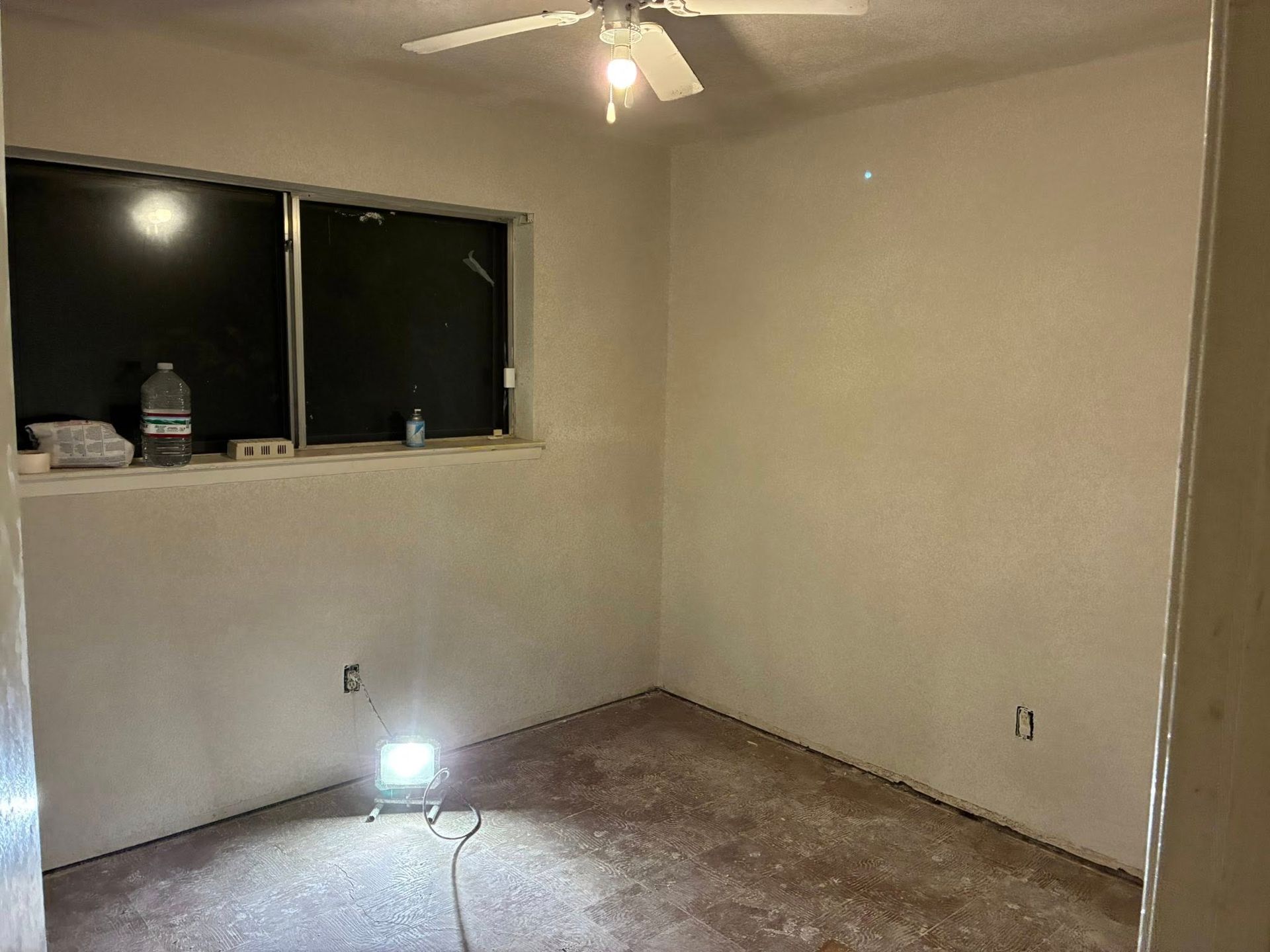 Empty beige room with carpet, window, ceiling fan, and a bright floor light near the wall.