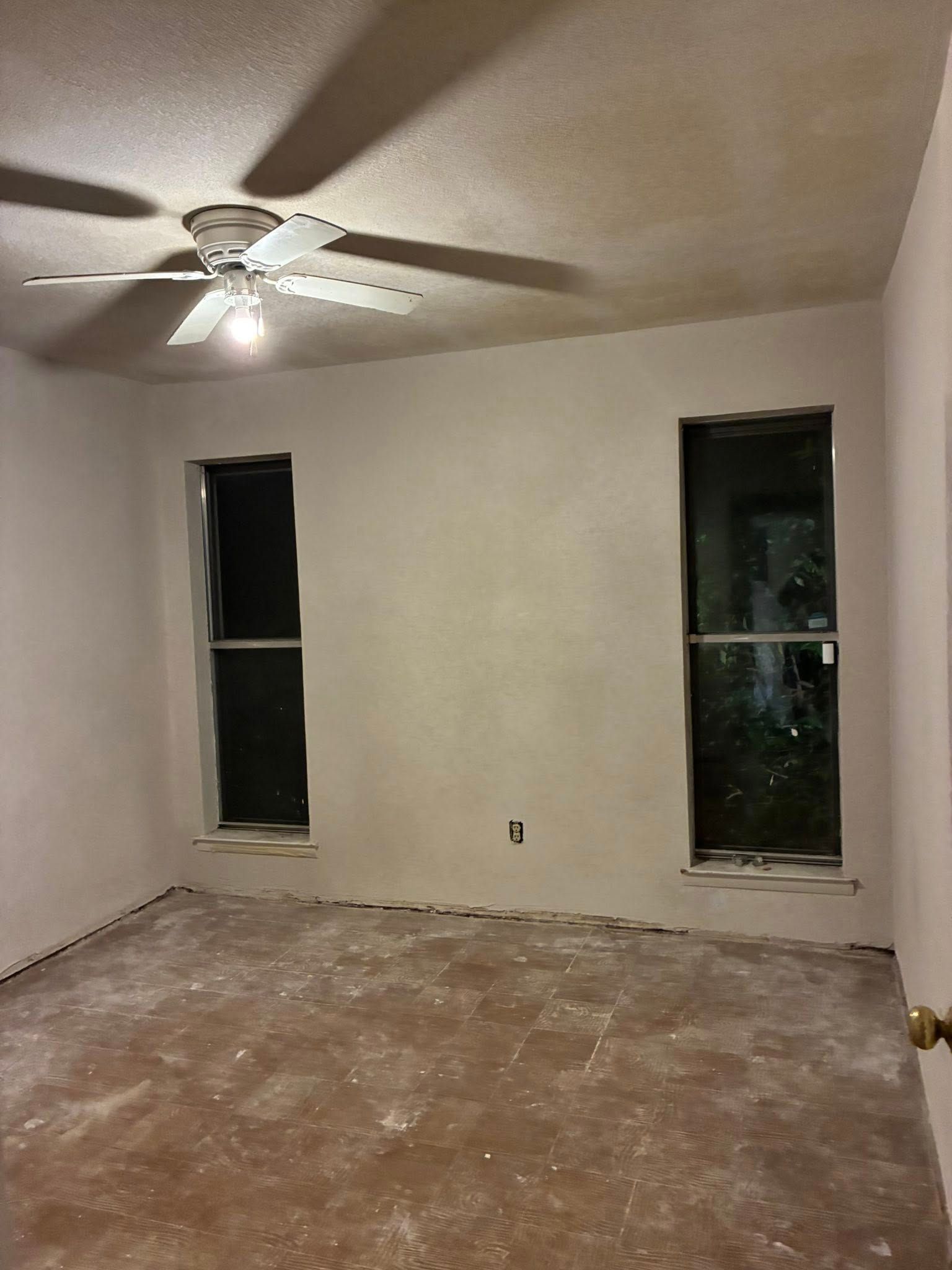 Empty beige bedroom with ceiling fan, two narrow windows, and worn carpet flooring.