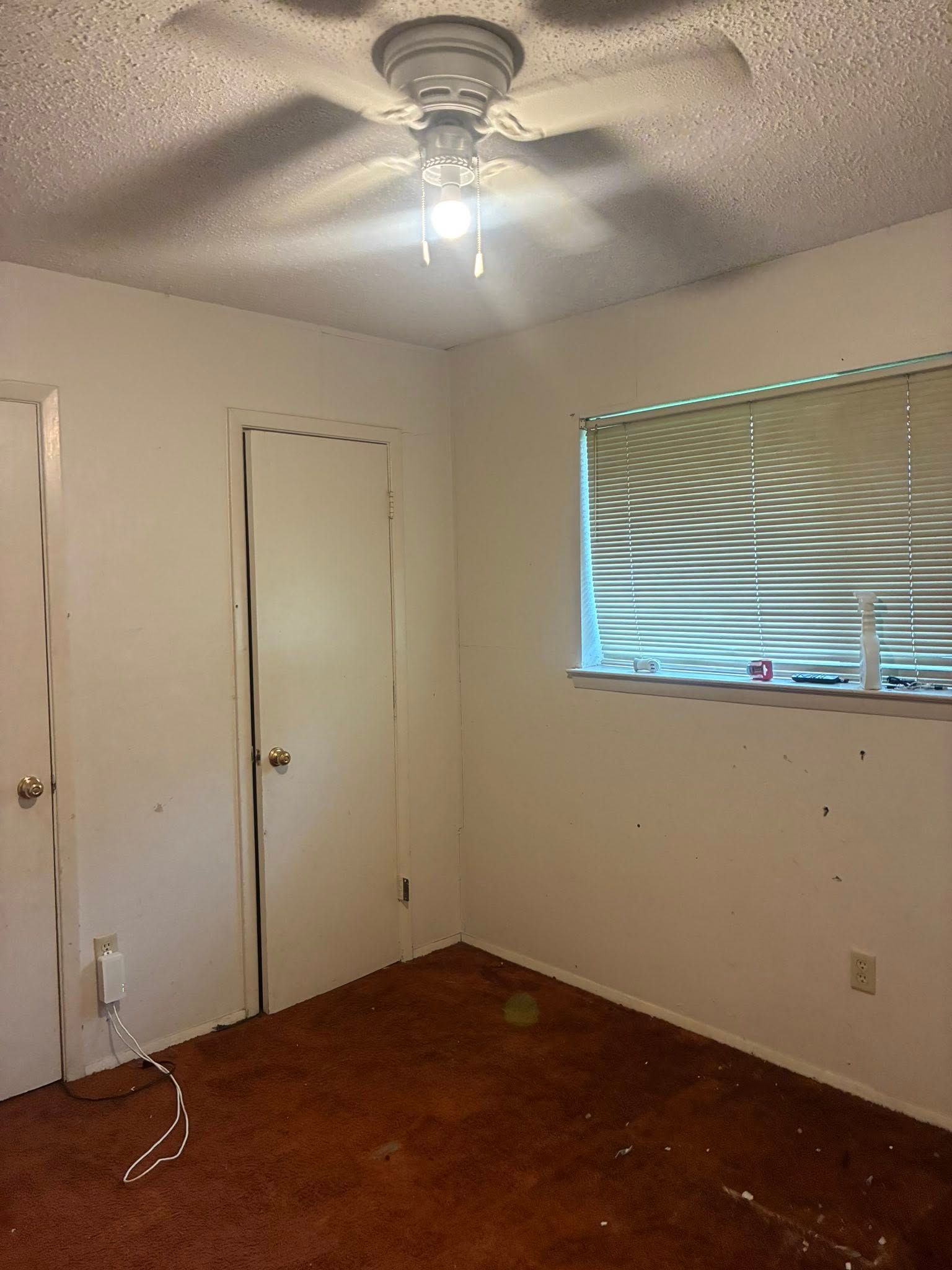 Empty bedroom with white walls, brown carpet, ceiling fan, closed door, and window with blinds.