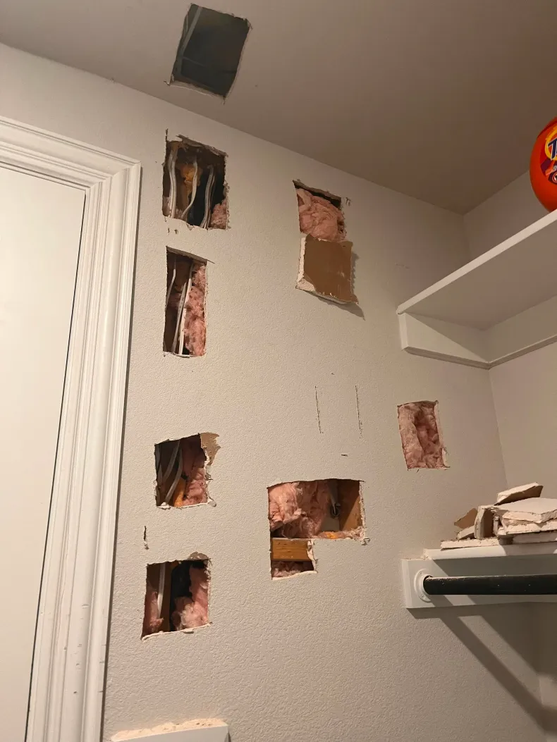 Damaged white wall with multiple holes exposing insulation, beside shelves and a ceiling vent