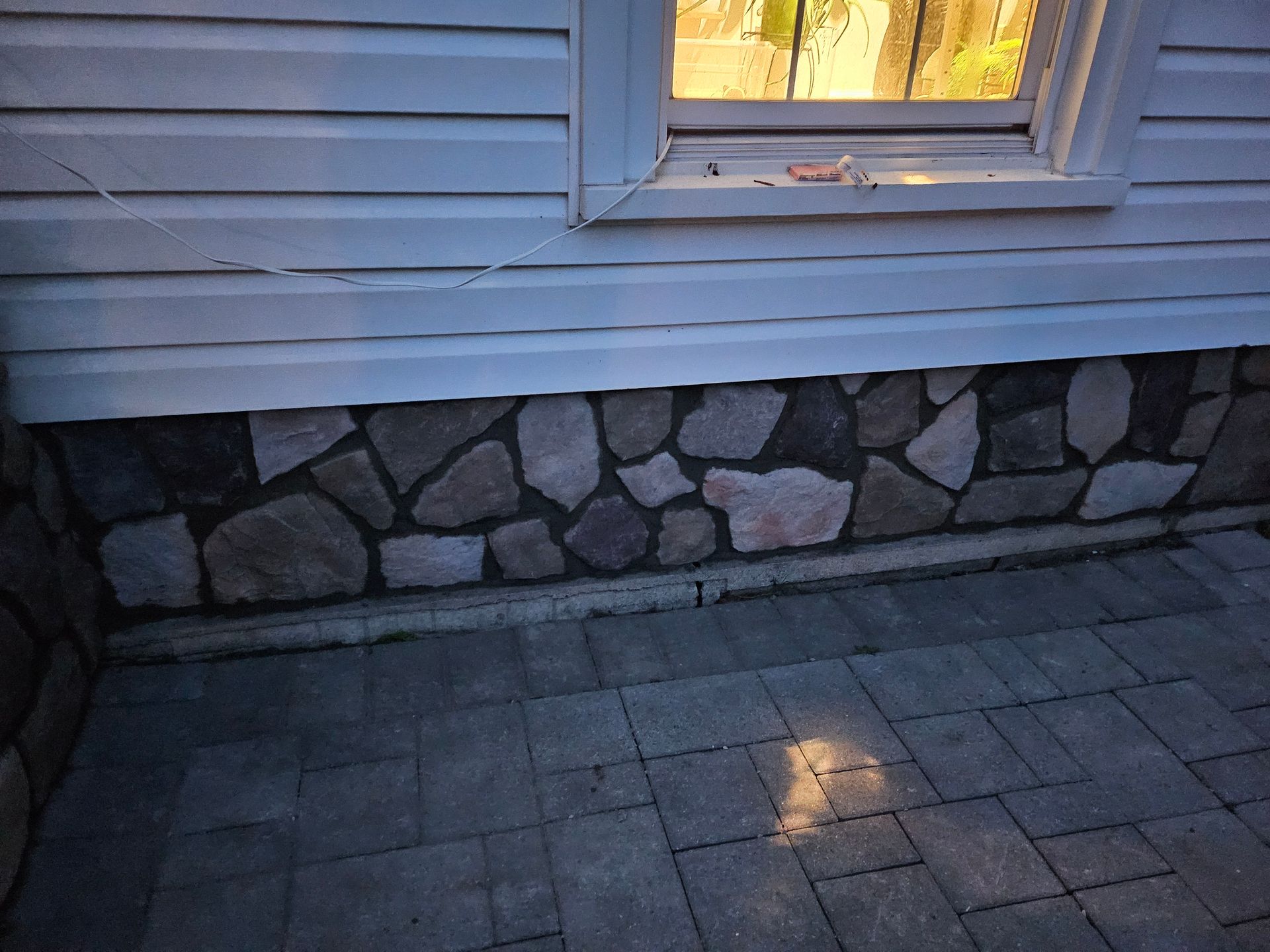 A house exterior showing a stone foundation below white horizontal siding, a window, and a gray paver patio floor.