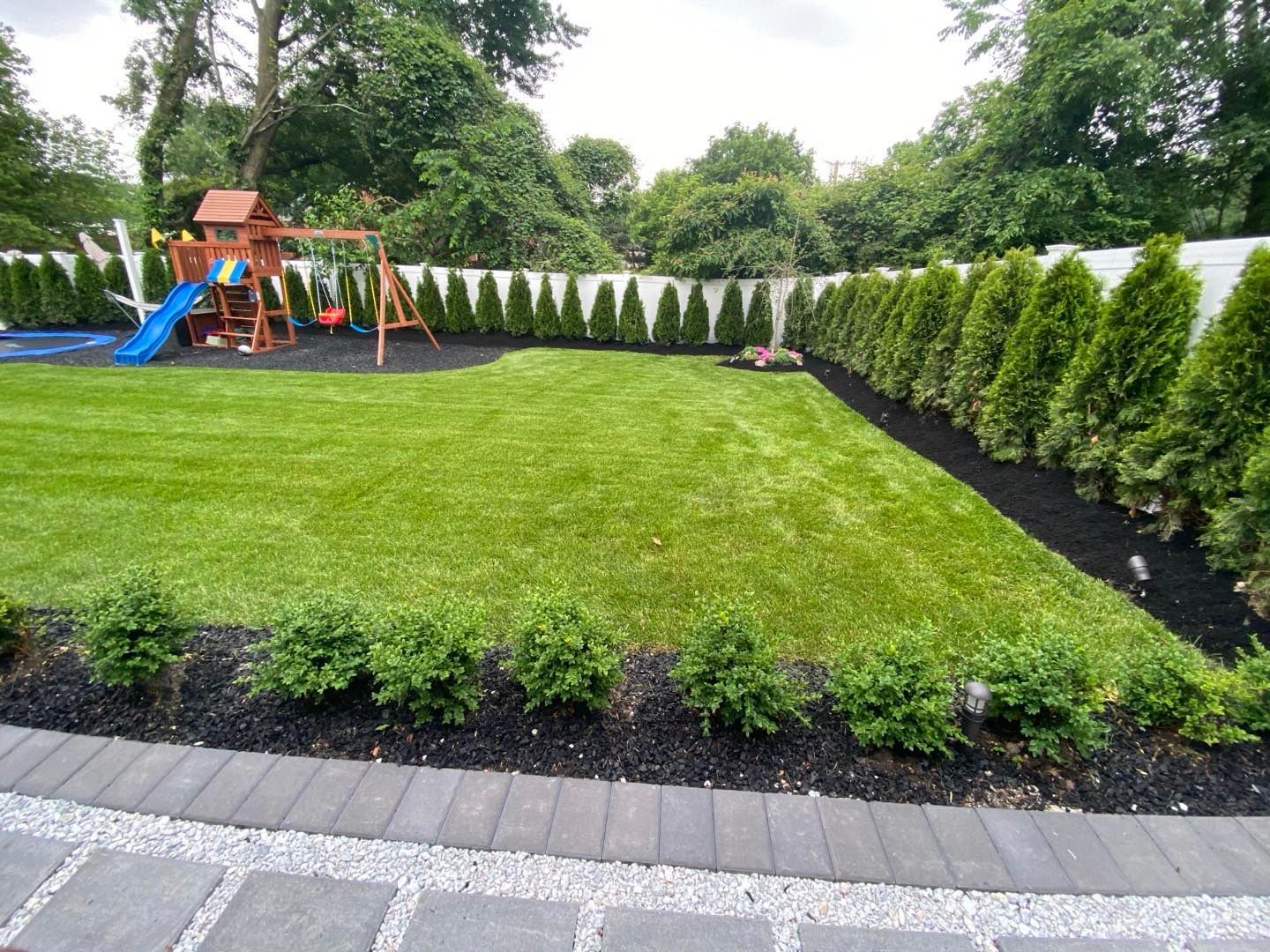 A landscaped backyard featuring a green lawn, a wooden playground set, and a row of evergreen trees along a white fence.