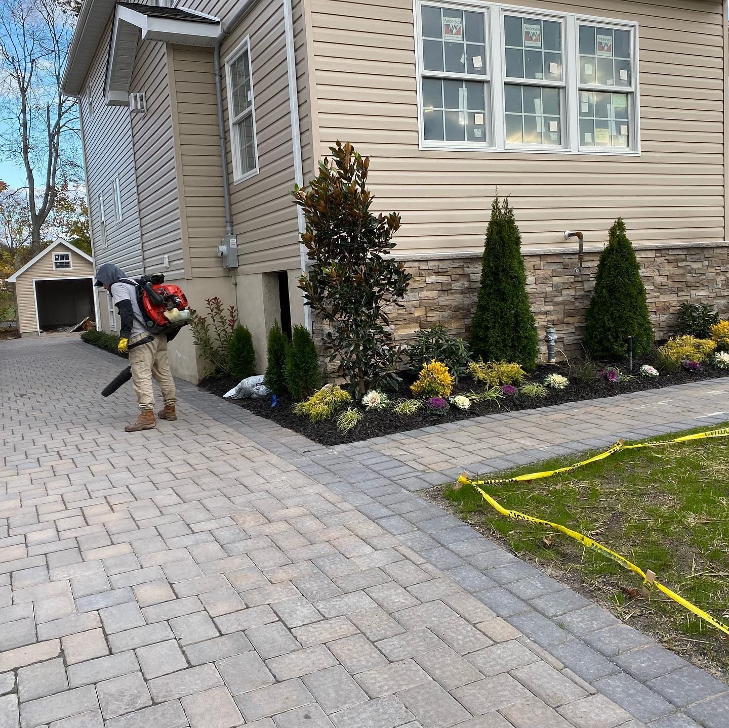 A person wearing protective gear uses a leaf blower to clear debris from a paved driveway beside a suburban home.