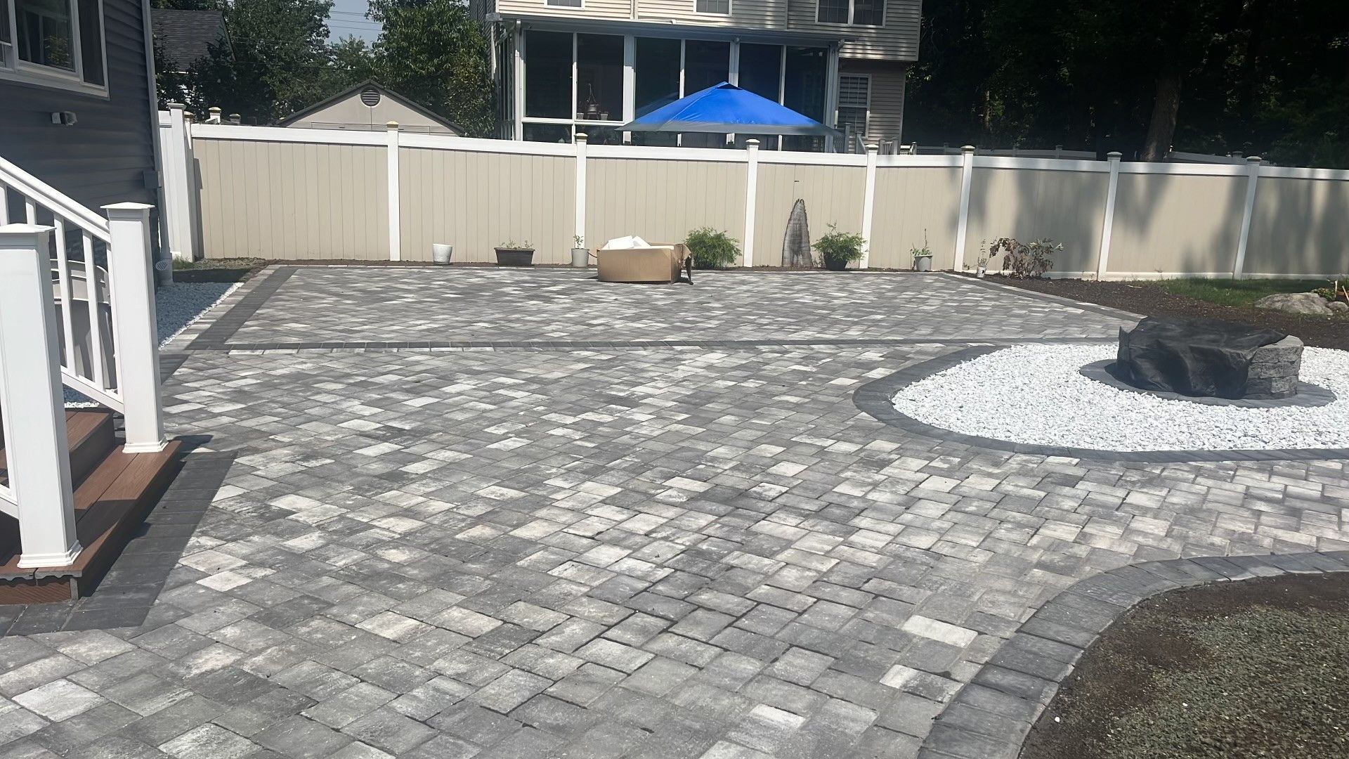 A backyard patio made of grey patterned pavers featuring a fire pit area with white stones, bordered by a beige fence.