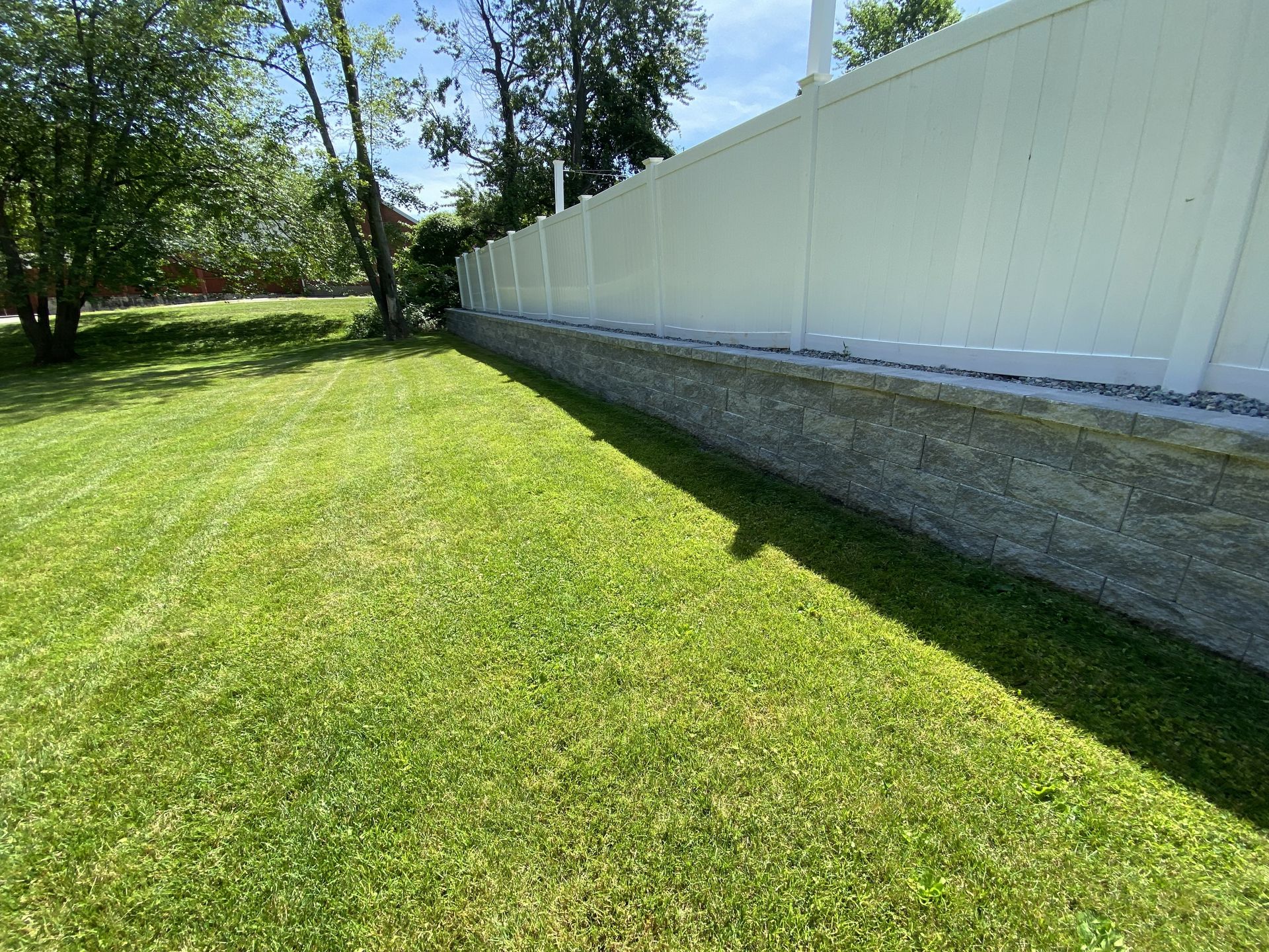A bright green lawn next to a low stone retaining wall topped with a white vinyl fence under a sunny blue sky.
