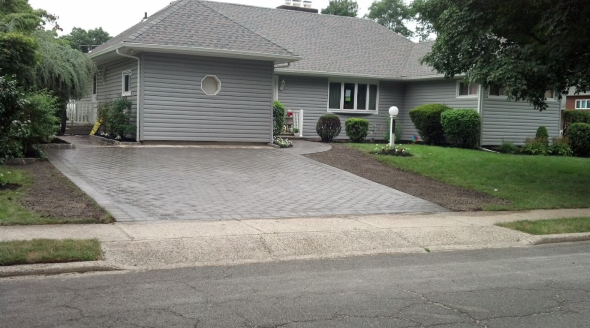 A gray house with a new light gray paver driveway and landscaped garden beds on both sides, viewed from the street.