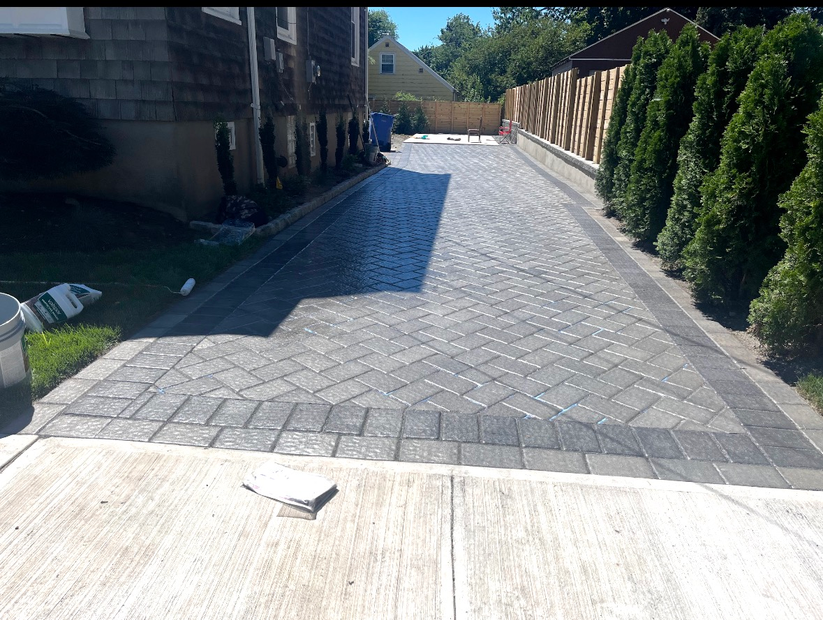 A paved driveway with a gray herringbone brick pattern, bordered by dark pavers, alongside a house and a row of shrubs.