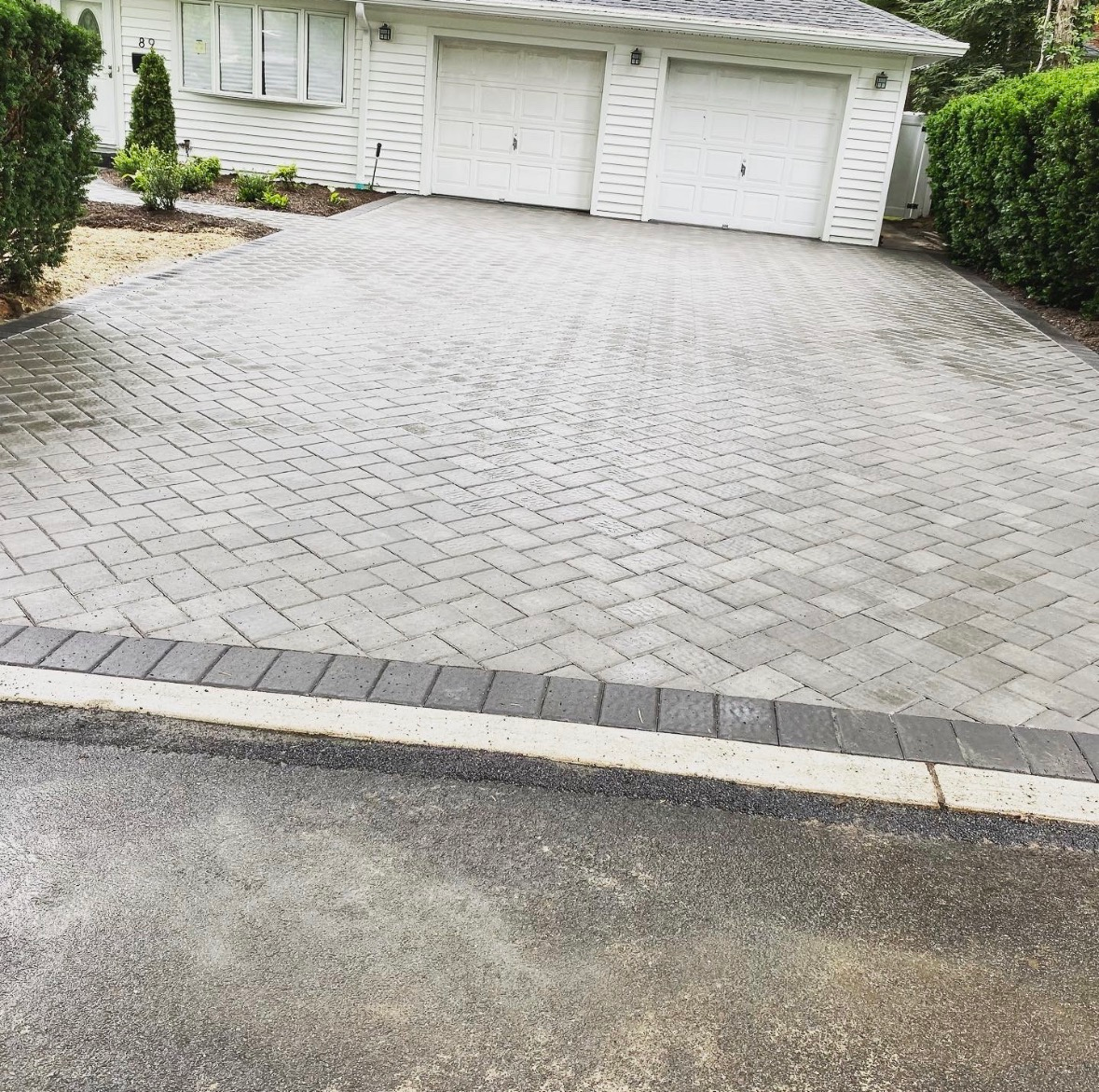 A gray interlocking paver driveway with a dark border leads to a white two-car garage at a suburban home.