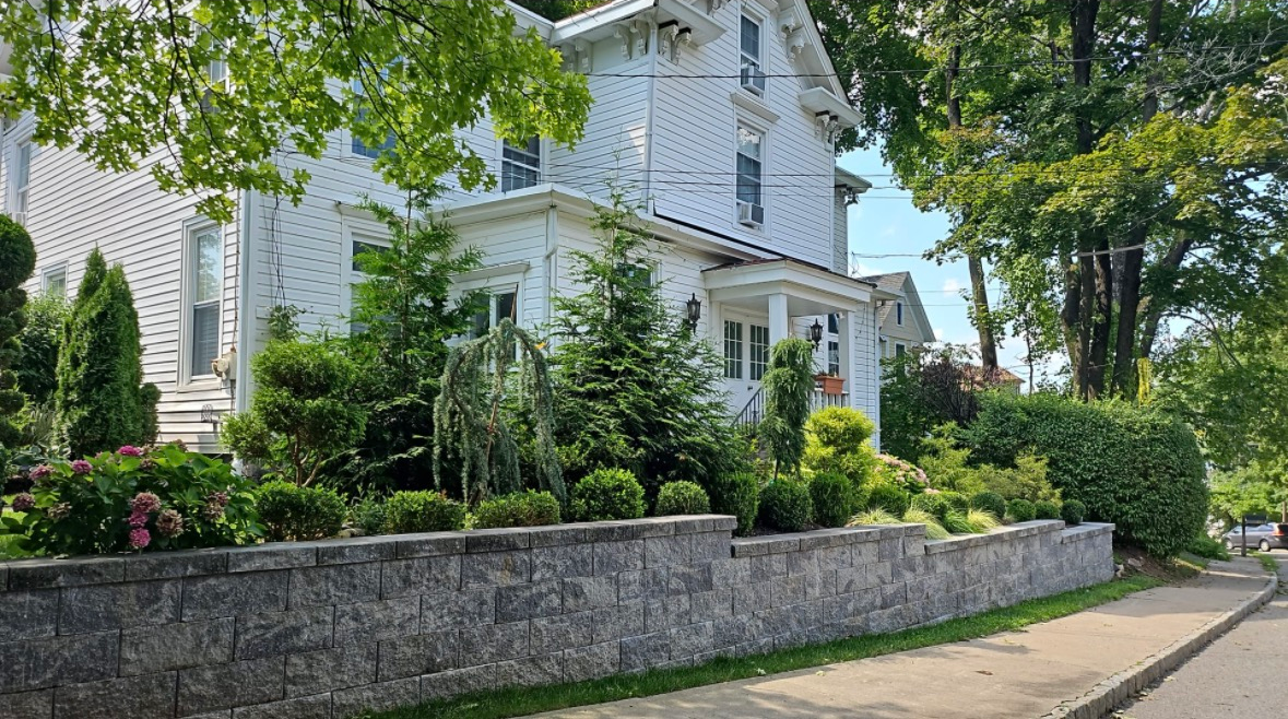 A large, white, multi-story house sits behind a stone retaining wall and lush landscaping on a sunny, tree-lined street.